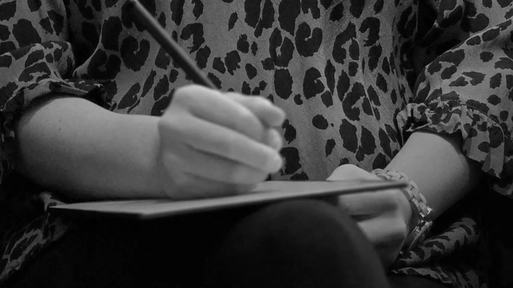 Henkilö kirjoittaa tai piirtää muistikirjaan, käden ja osan ranteesta näkyvissä, pukeutuneena eläinprinttikuvioiseen vaatteeseen.