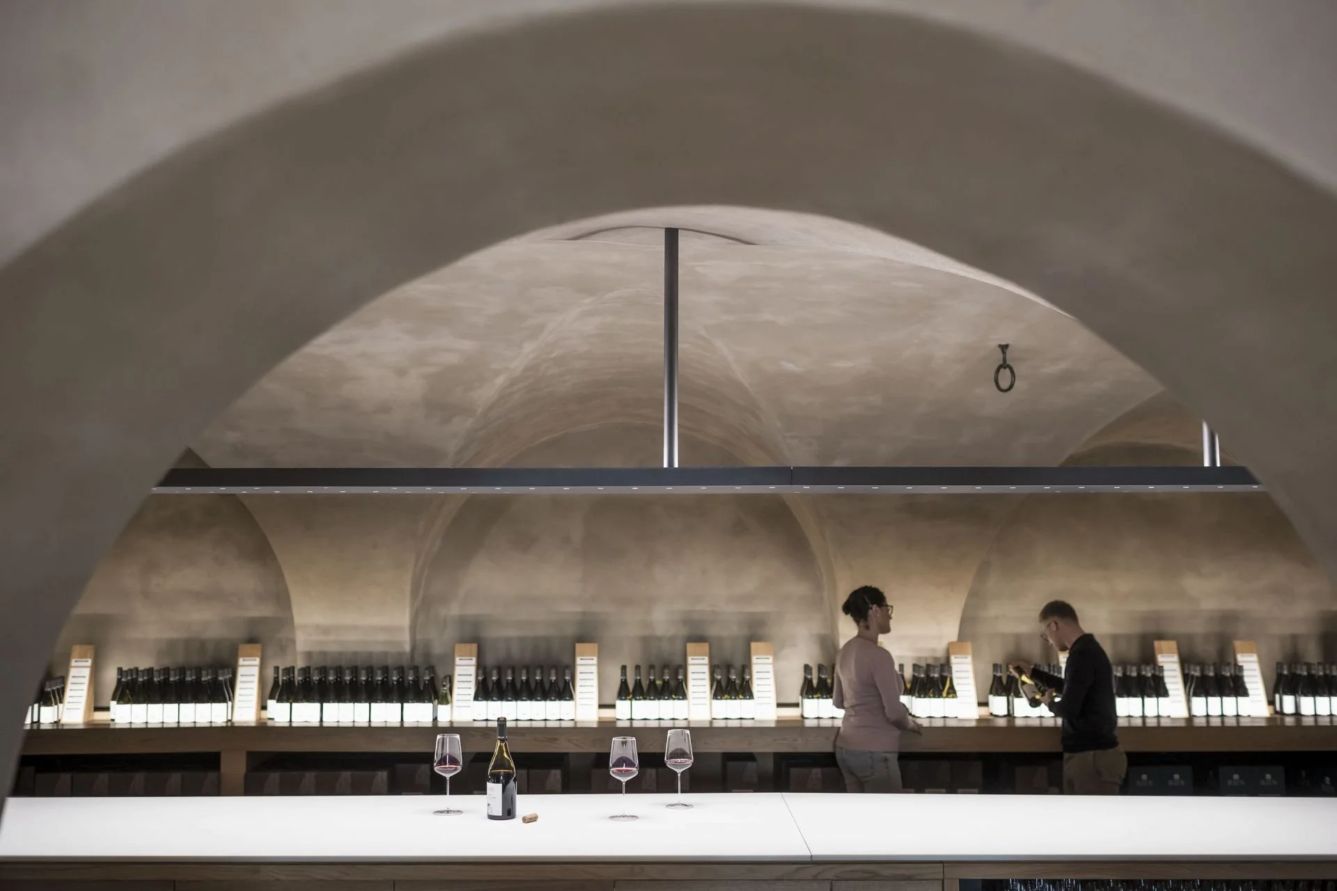 Interior of a wine cellar or tasting room with arched ceilings, a long counter with wine bottles, and two people conversing, with wine glasses on the counter.