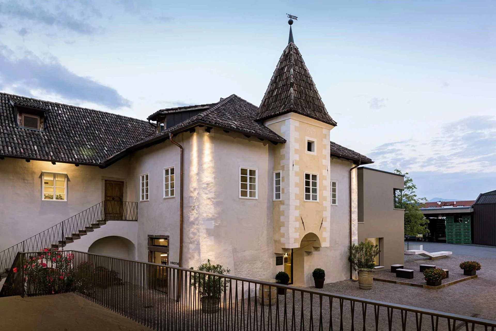 A European-style building with a tower, tiled roof, and painted white walls, surrounded by potted plants and outdoor seating.