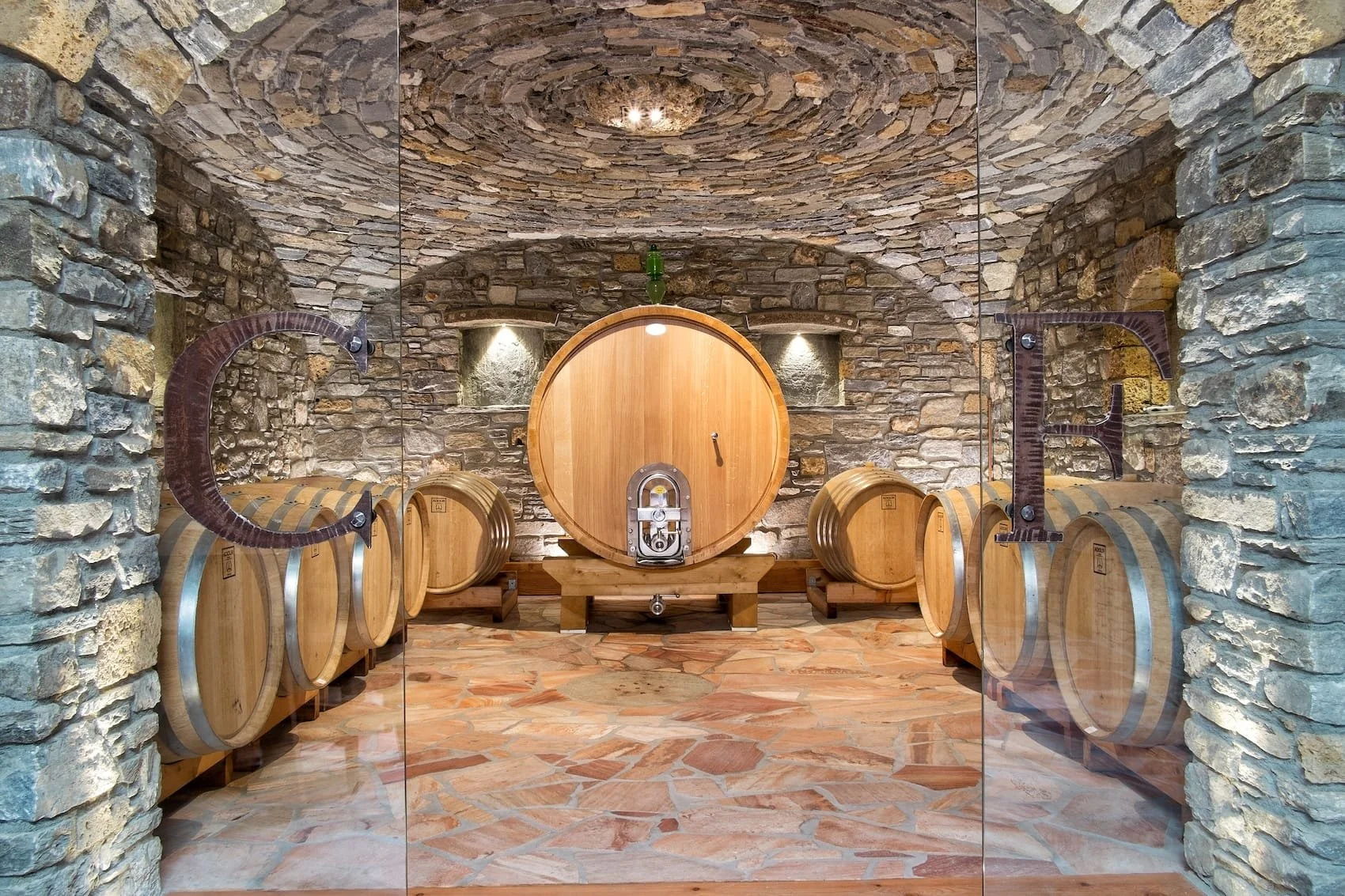 A wine cellar with stone walls and ceiling, wooden barrels lined along both sides, and a large central wine barrel with a tap, glass door, and metal fittings, seen through a glass wall with metal fixtures.