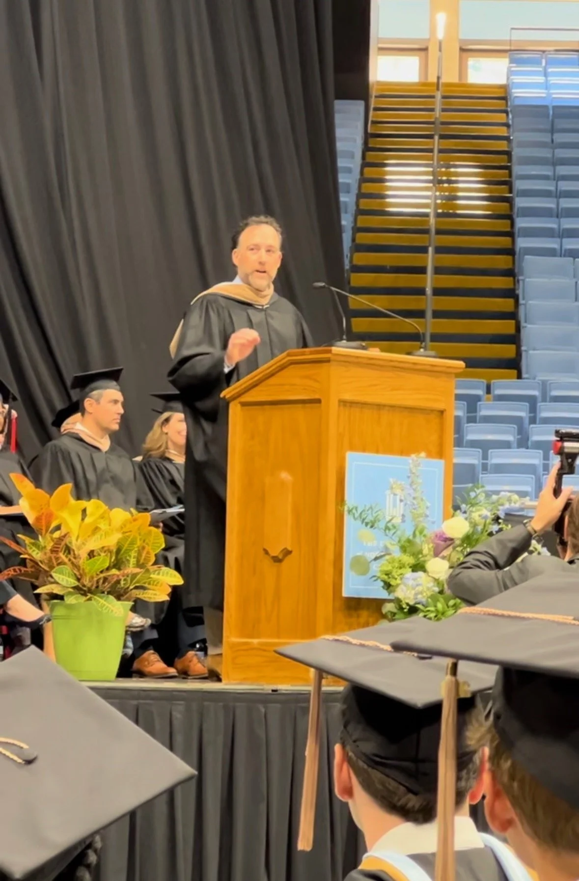 A man giving a speech at a graduation ceremony, standing behind a wooden podium decorated with flowers, with students in graduation caps and gowns seated nearby and an audience in the foreground.