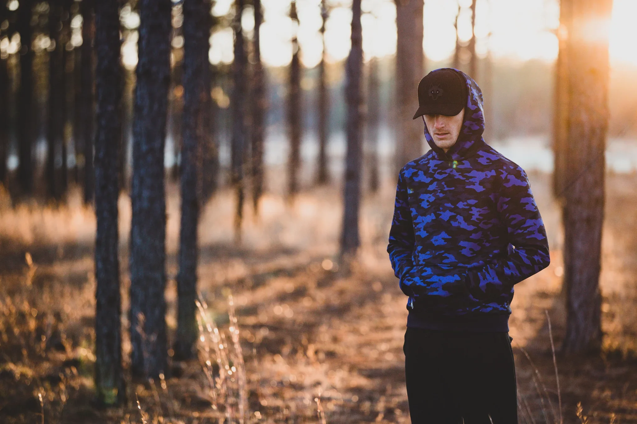 A man in a blue camouflage hoodie and black cap stands in a forest during sunset.