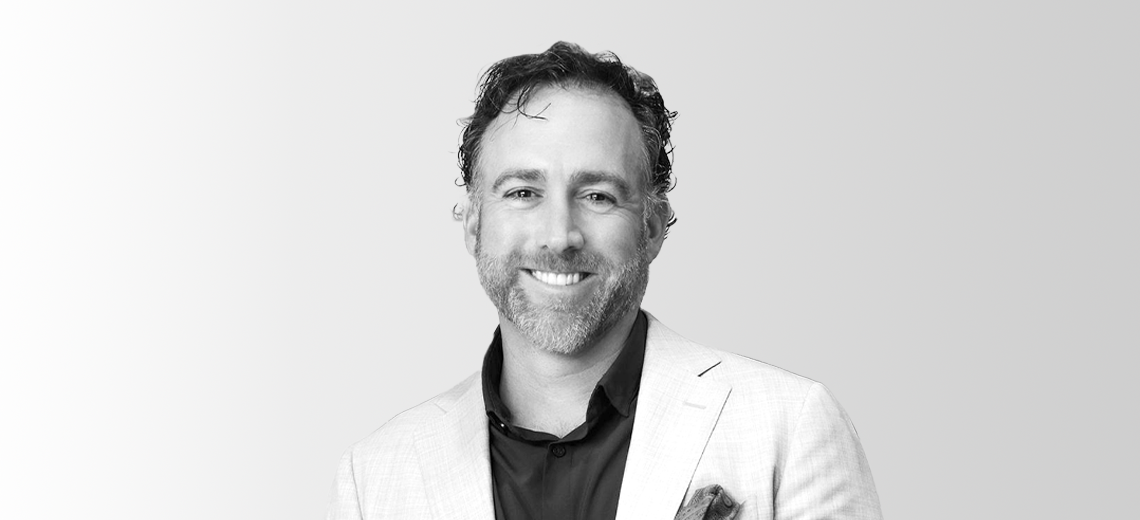 Black and white portrait of a smiling man with short curly hair and beard, wearing a light-colored blazer and dark shirt.