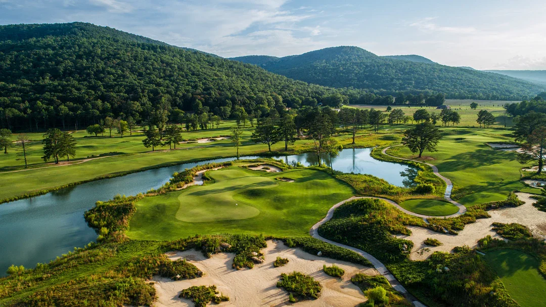 Aerial view of a golf course with green fairways, water hazards, sand traps, and trees, surrounded by rolling hills and mountains.