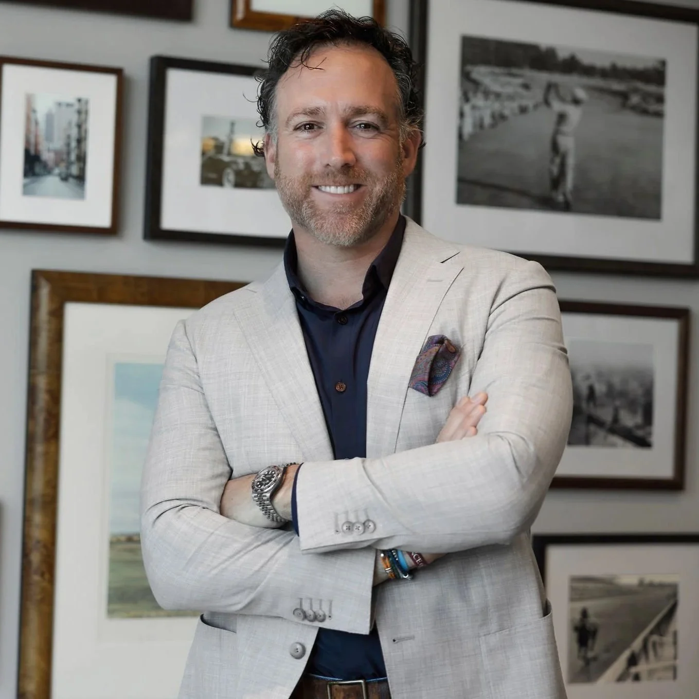 A man standing with arms crossed, smiling, in front of a wall with multiple framed black-and-white photographs.