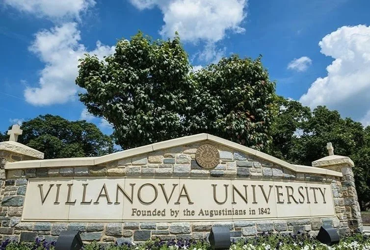 Villanova University entrance sign with stone wall and trees in the background, under a blue sky with clouds.