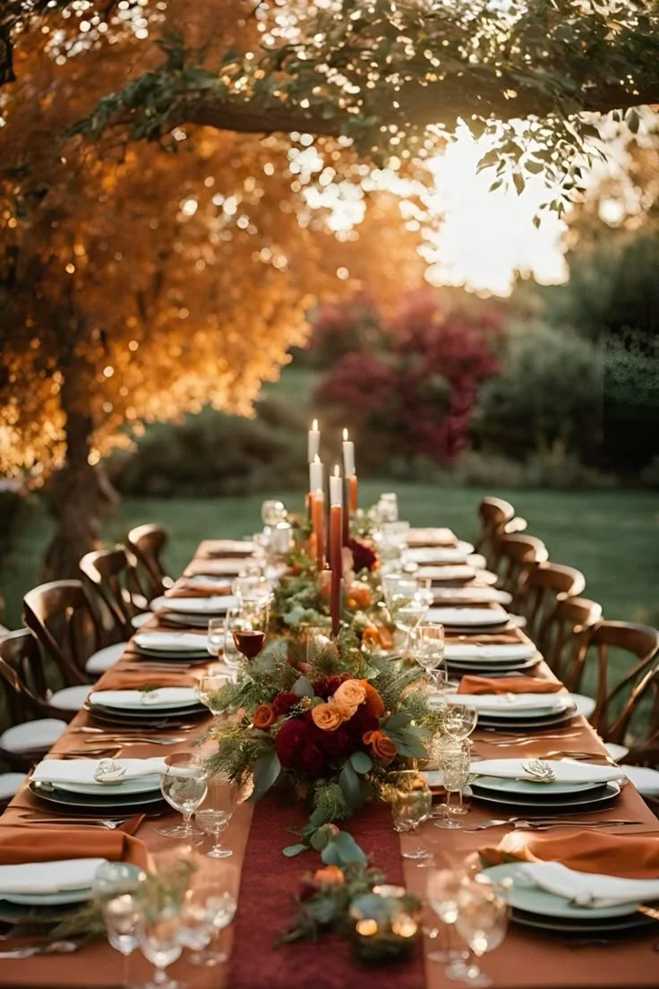 Outdoor dining table set for a celebration with floral centerpieces and candles, surrounded by trees with autumn leaves.