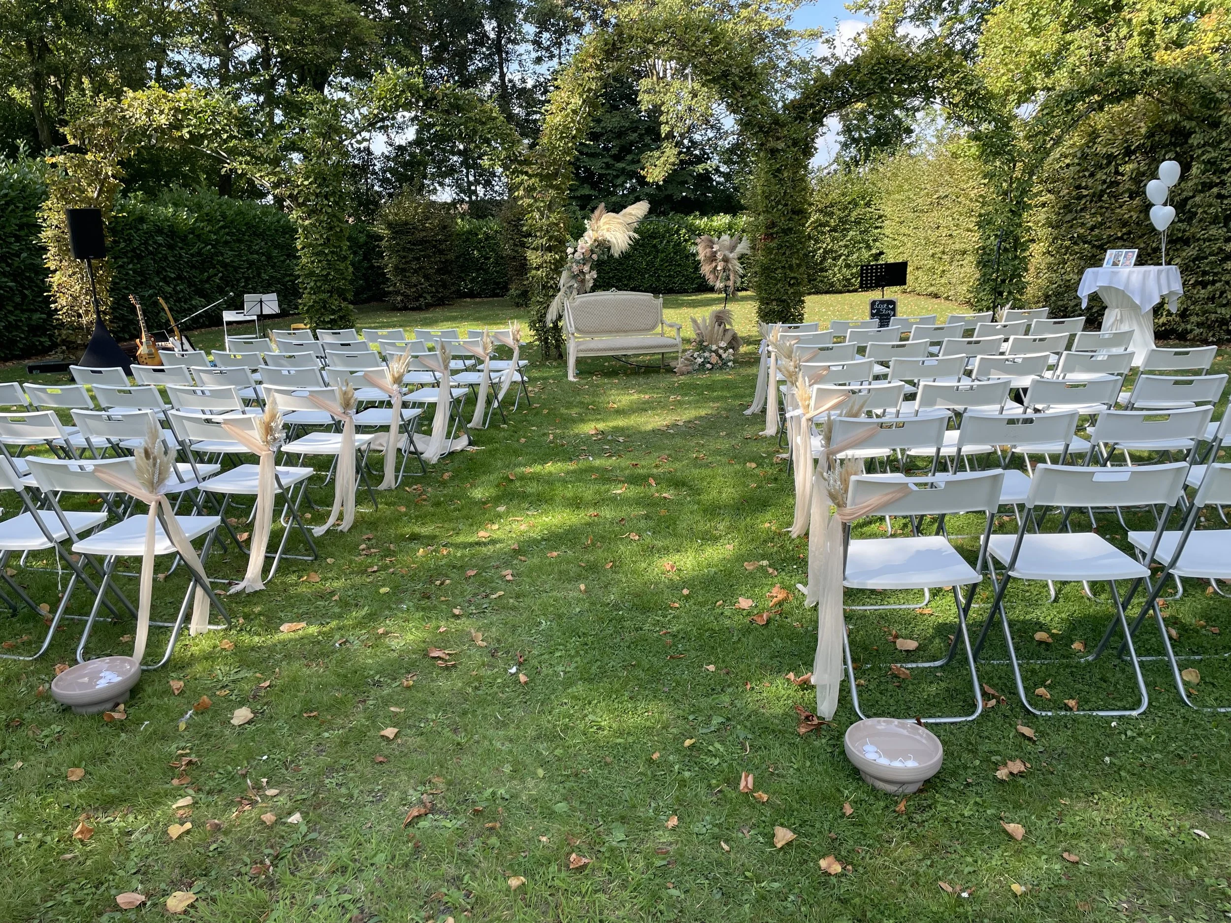 Outdoor wedding ceremony setup with white folding chairs decorated with beige fabric and dried floral arrangements on a grassy lawn, facing a white vintage loveseat and decorated arch, surrounded by trees and greenery.