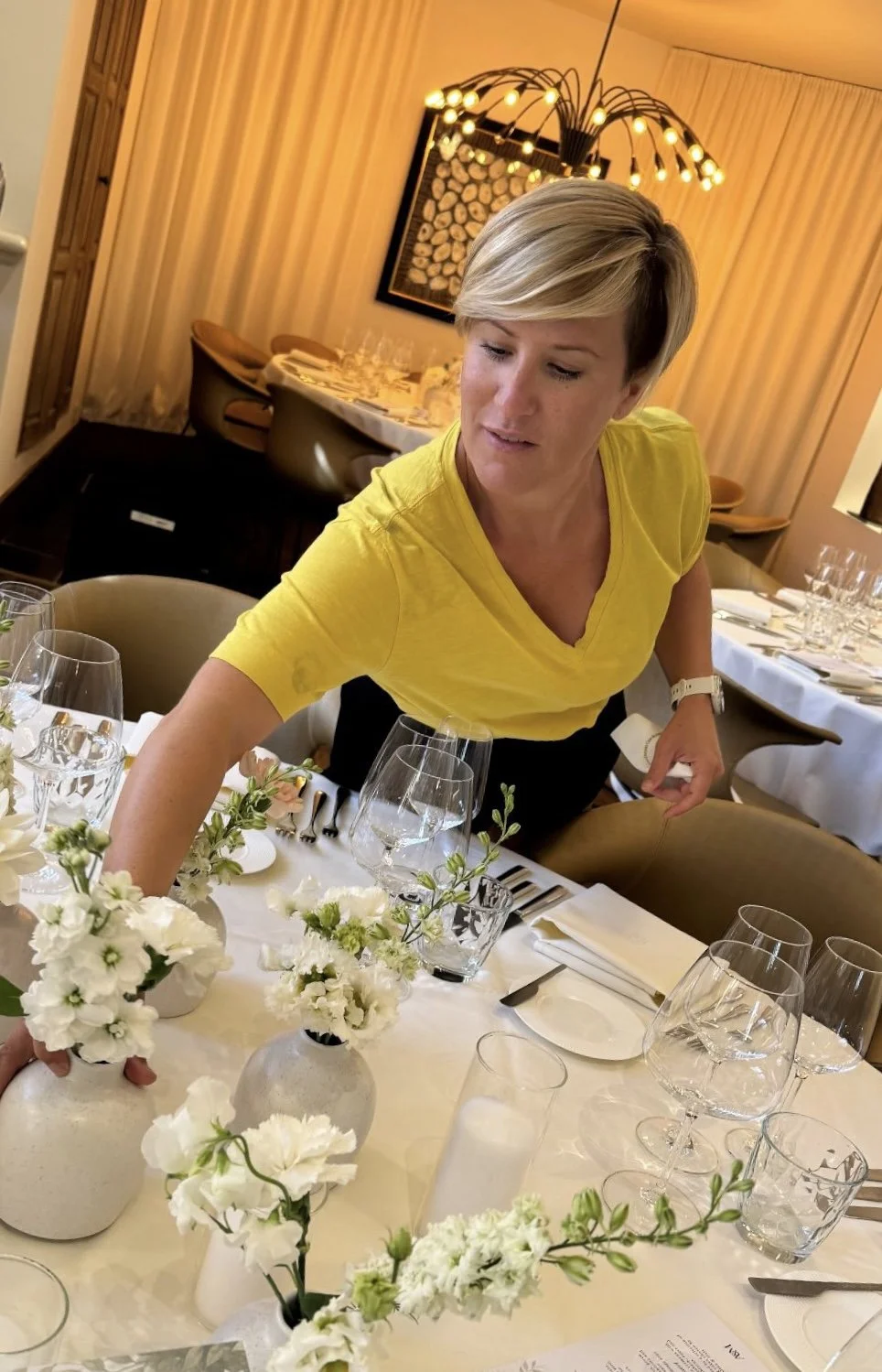A woman in a yellow shirt setting a dining table with white flowers and tableware in a nicely decorated restaurant.