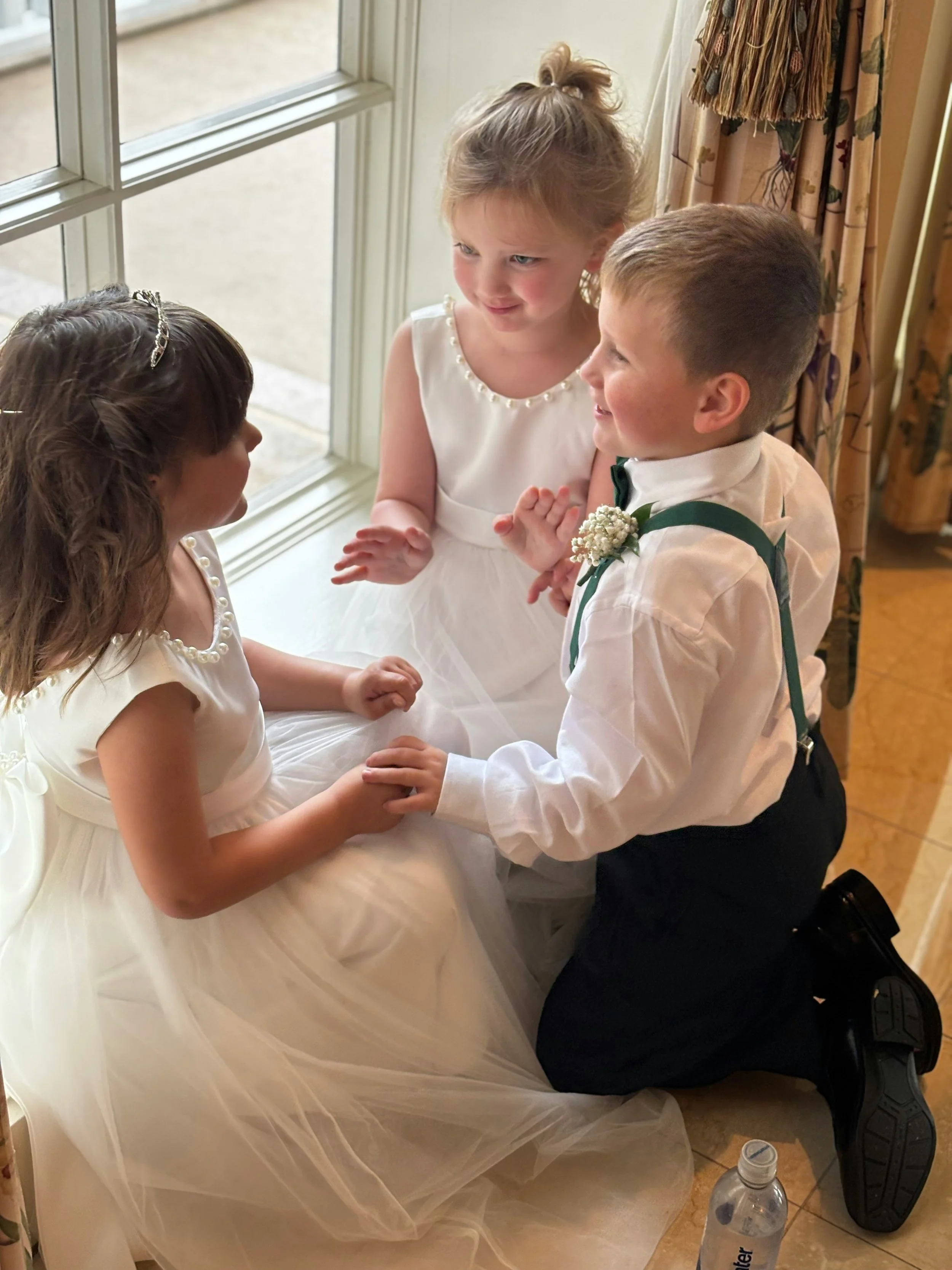 A young boy and girl holding hands and smiling at each other, dressed in formal wedding attire, with two young girls sitting by the window in their wedding dresses, in a room with floral curtains and a water bottle on the floor.