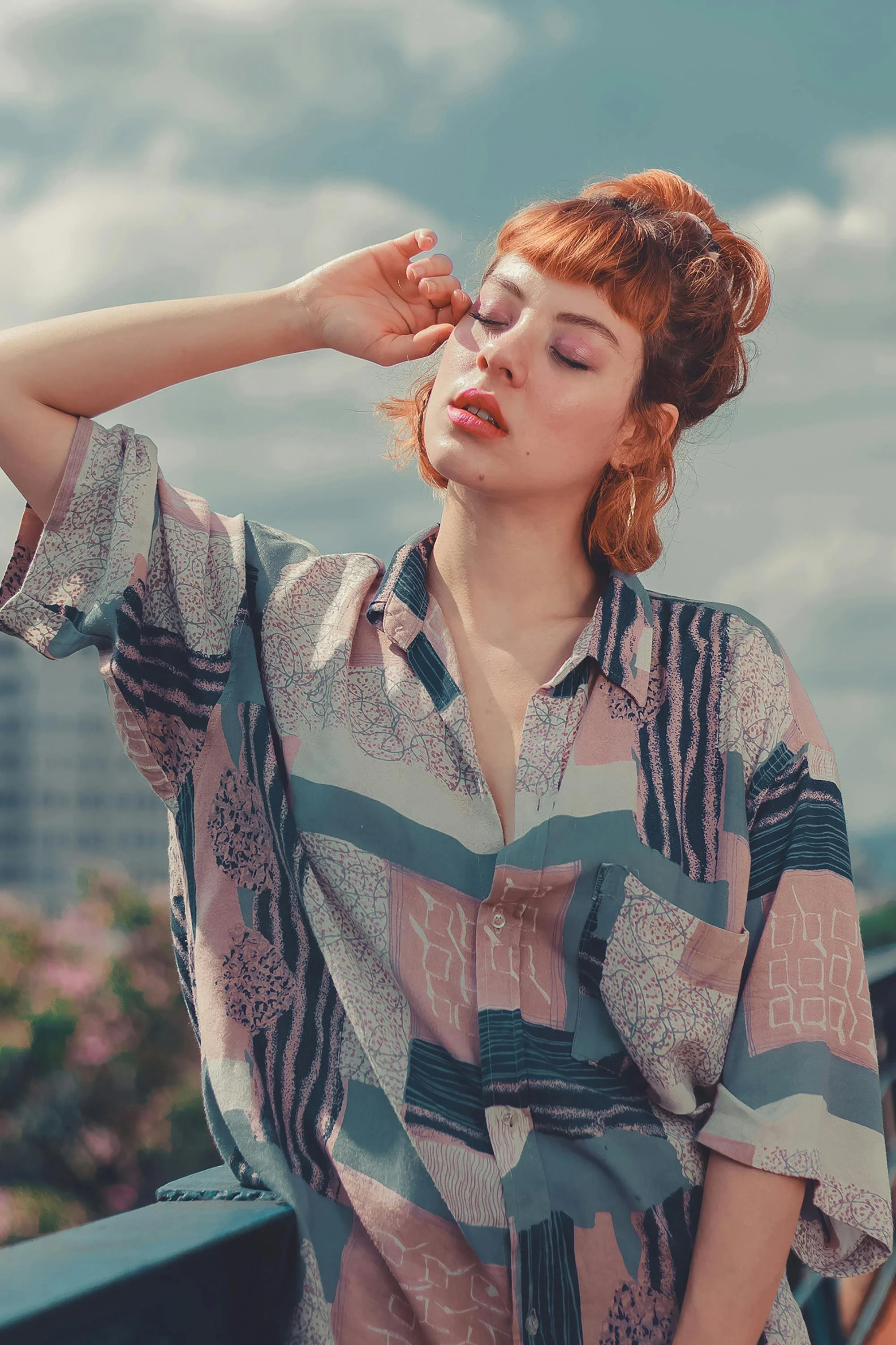 A red-haired woman in a multicolored patterned shirt is outdoors with a cloudy sky in the background. She has her eyes closed and one hand near her temple, appearing relaxed or contemplative.