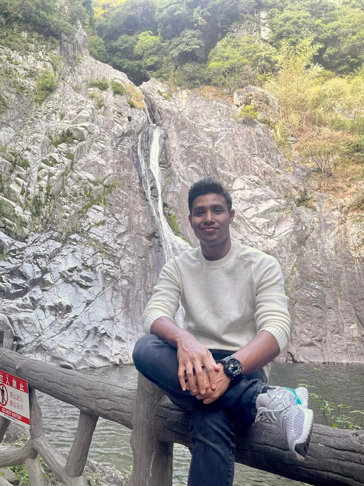 A young man sitting on a log in front of a mountain with a waterfall coming down the rocks, surrounded by green trees.