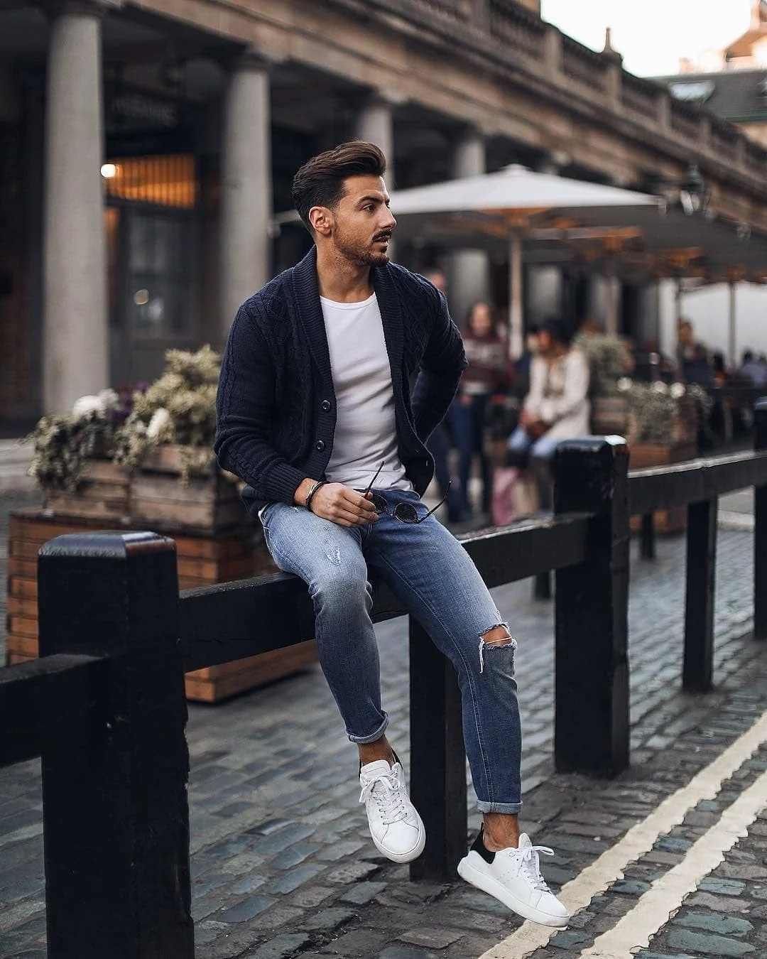 A young man in casual clothing sitting on a black wooden bench near a restaurant patio on a cobblestone street, holding sunglasses, with other people in the background.