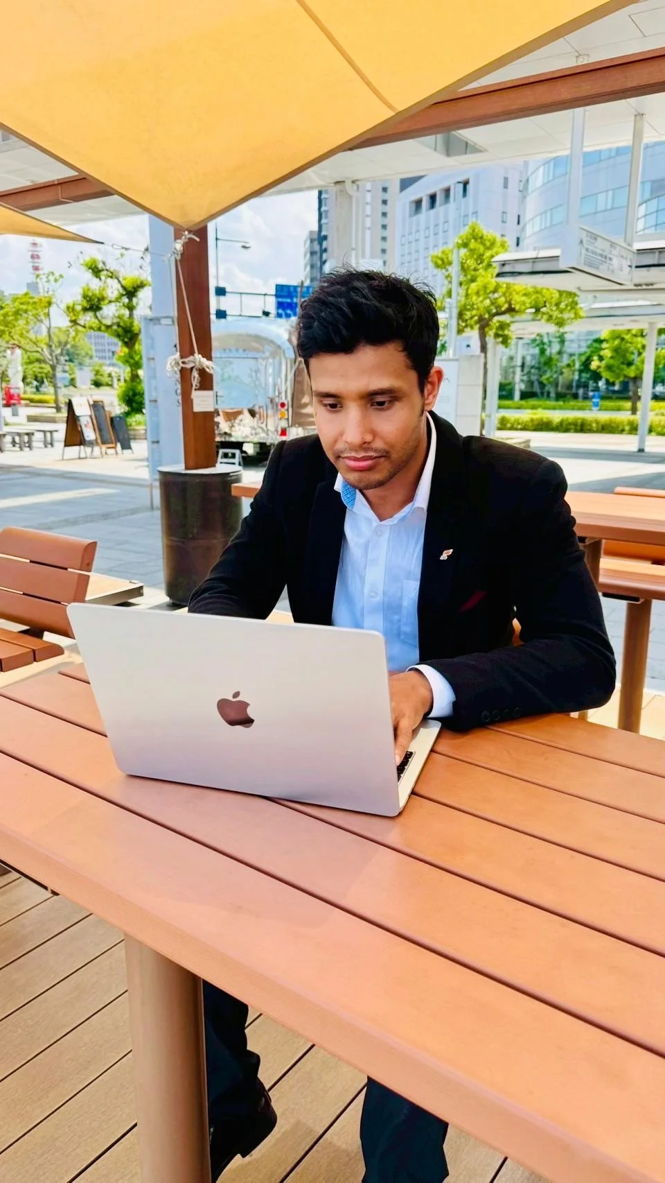 A man in a black suit jacket and white shirt sitting at a wooden outdoor table, working on a silver MacBook in an urban setting with trees, modern buildings, and a pedestrian walkway.