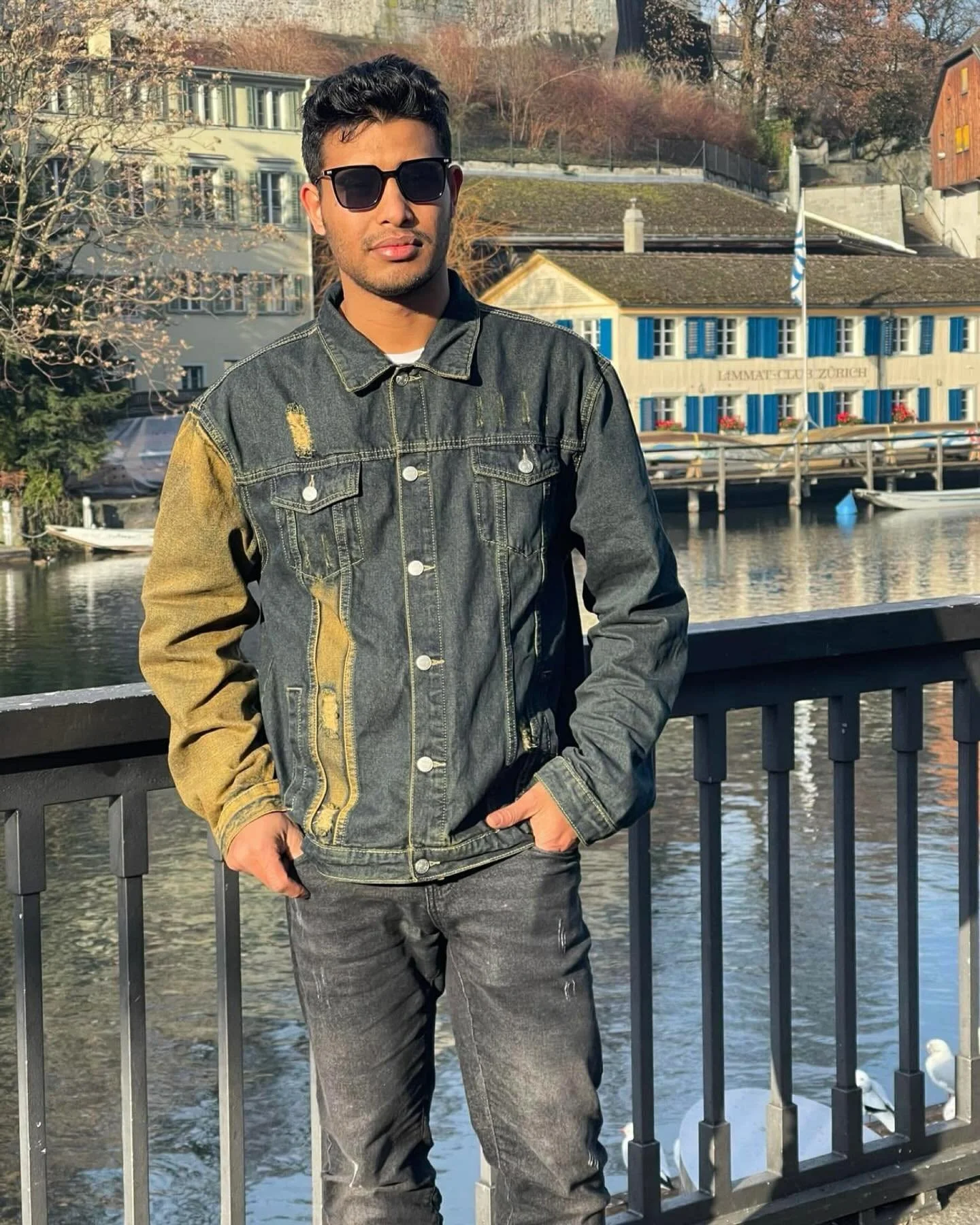 A young man wearing sunglasses and a distressed denim jacket stands by a waterway with buildings and trees in the background.