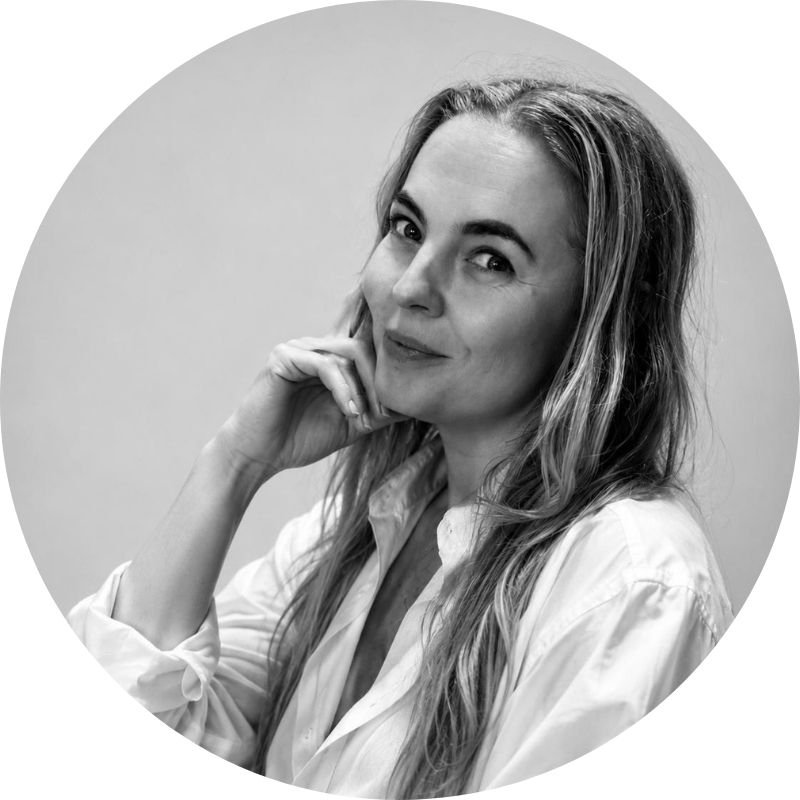 Black and white portrait of Dr Lisa Corbett with long wavy hair, wearing a white shirt, looking at the camera with her hand near her chin, smiling subtly.