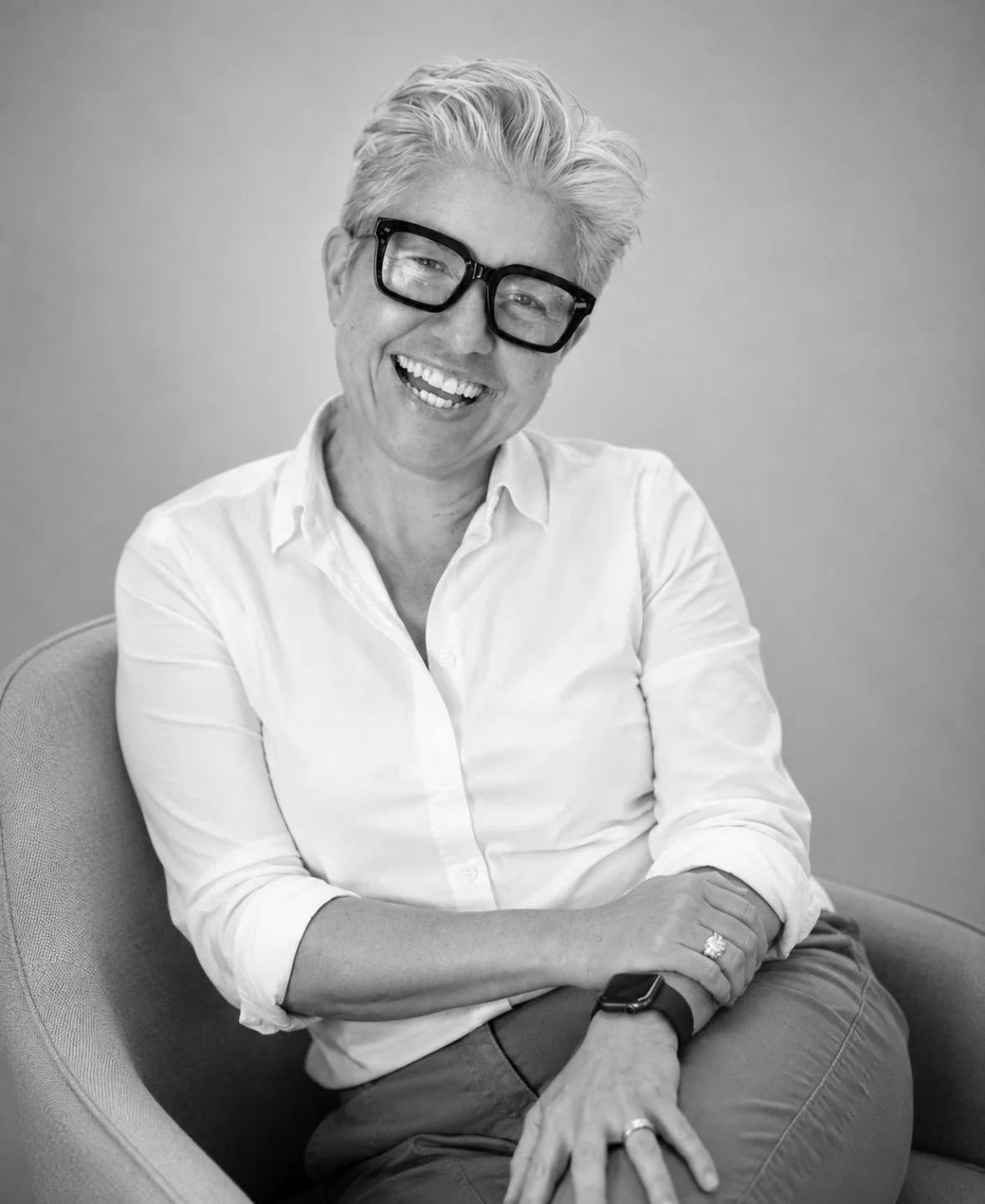 A smiling Dr Tanya Petersen with short, styled gray hair wearing glasses and a white button-up shirt, sitting in a chair against a plain background.