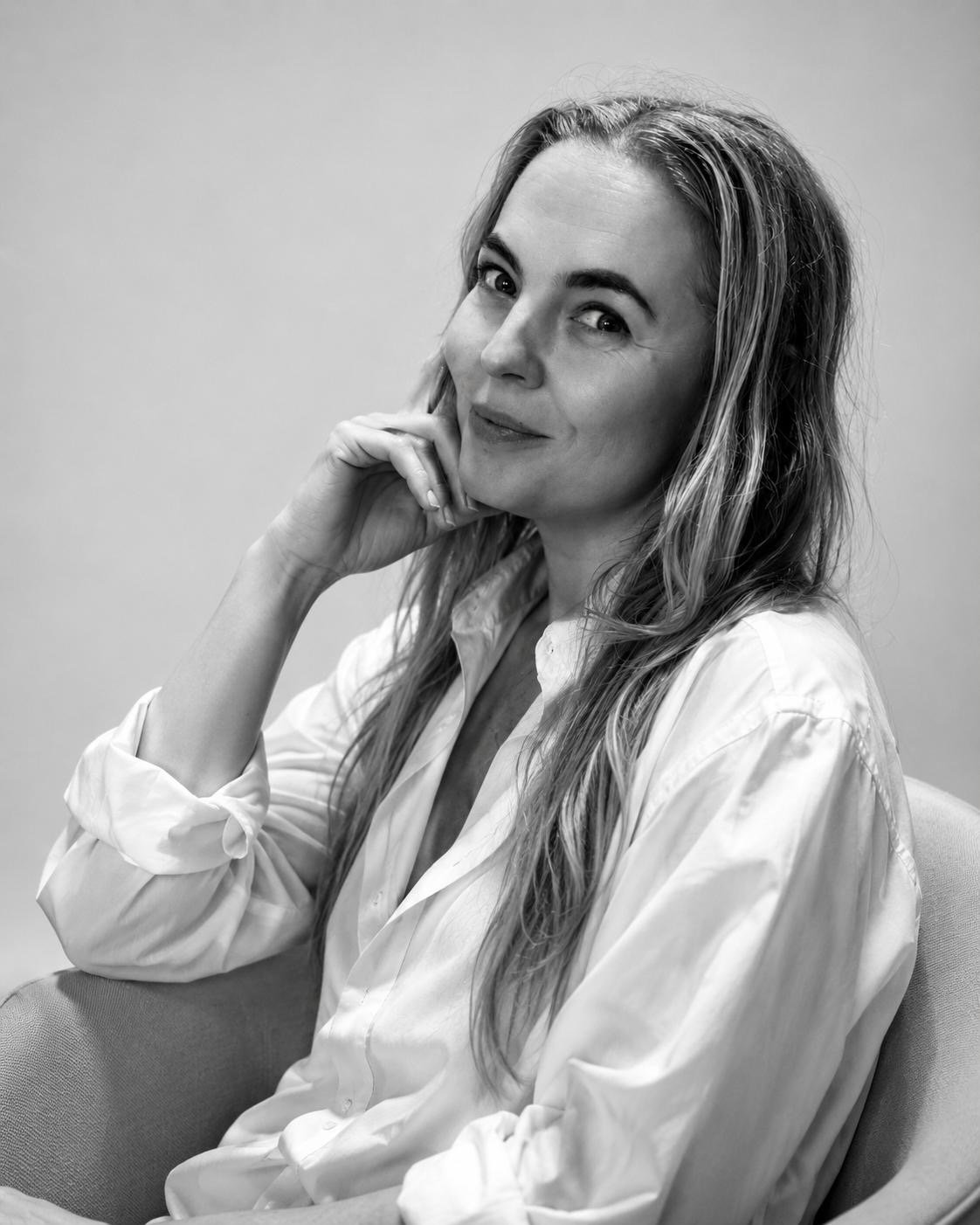 Black and white photo of Dr Lisa Corbett with long hair, wearing a white shirt, sitting on a chair, smiling gently, with one hand resting on her cheek against a plain background.