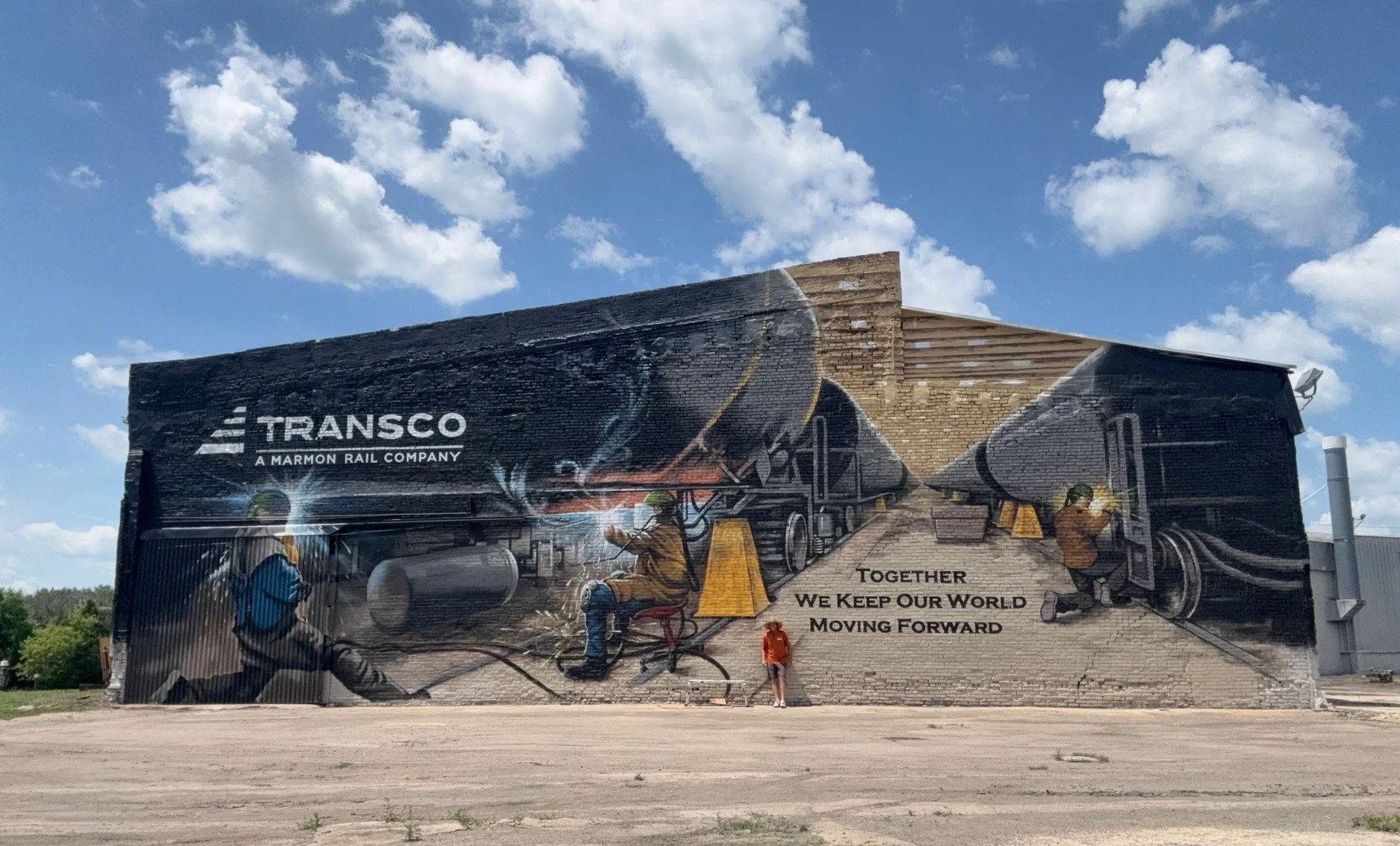 A large mural on a brick building depicting workers welding and maintaining train tracks, with the words "Transco A Marmon Rail Company" and a slogan "Together We Keep Our World Moving Forward". There are clouds in the sky above.