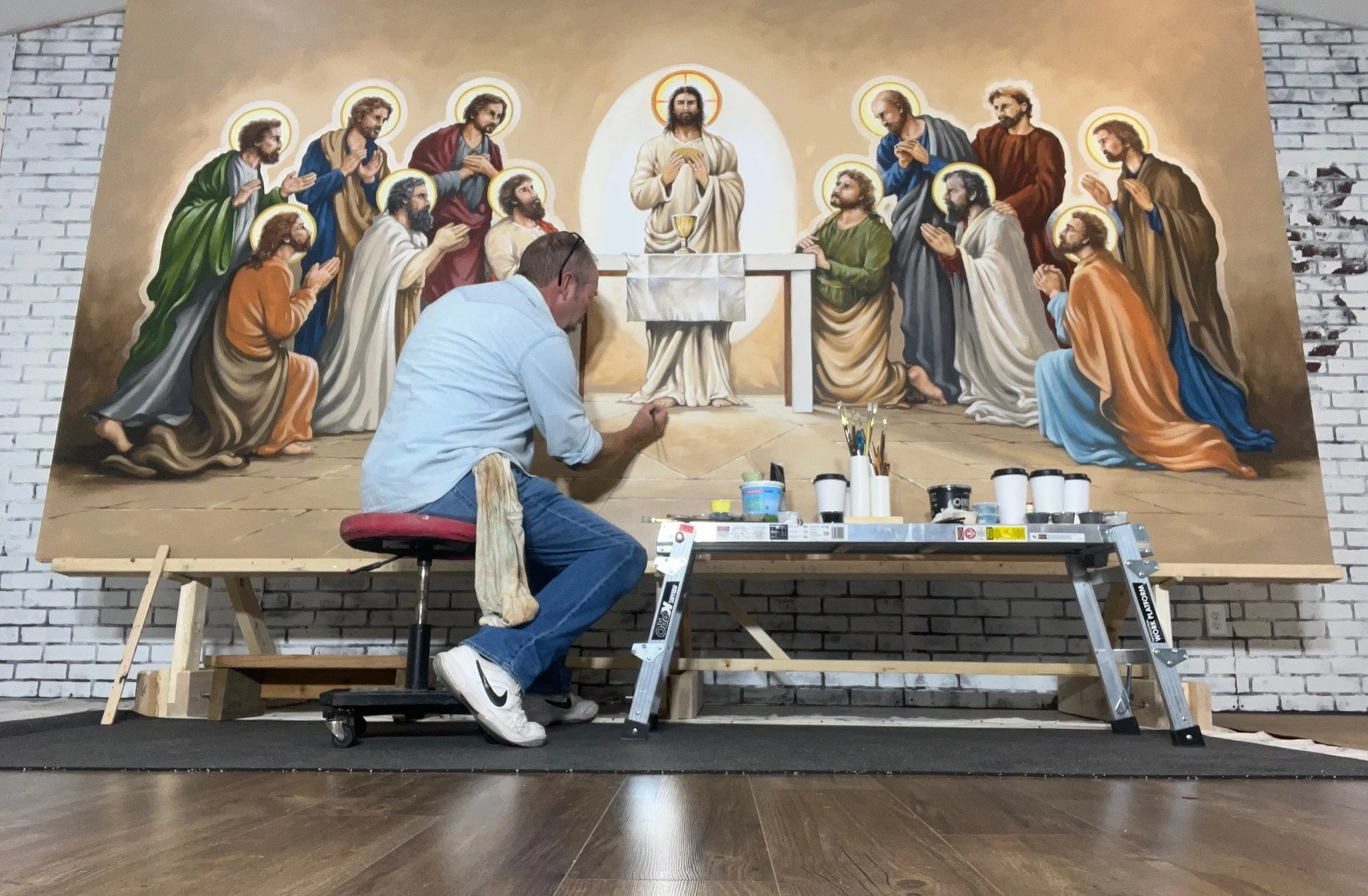 Artist painting a large religious mural depicting Jesus Christ and the apostles in an indoor studio, with art supplies on a table in front.