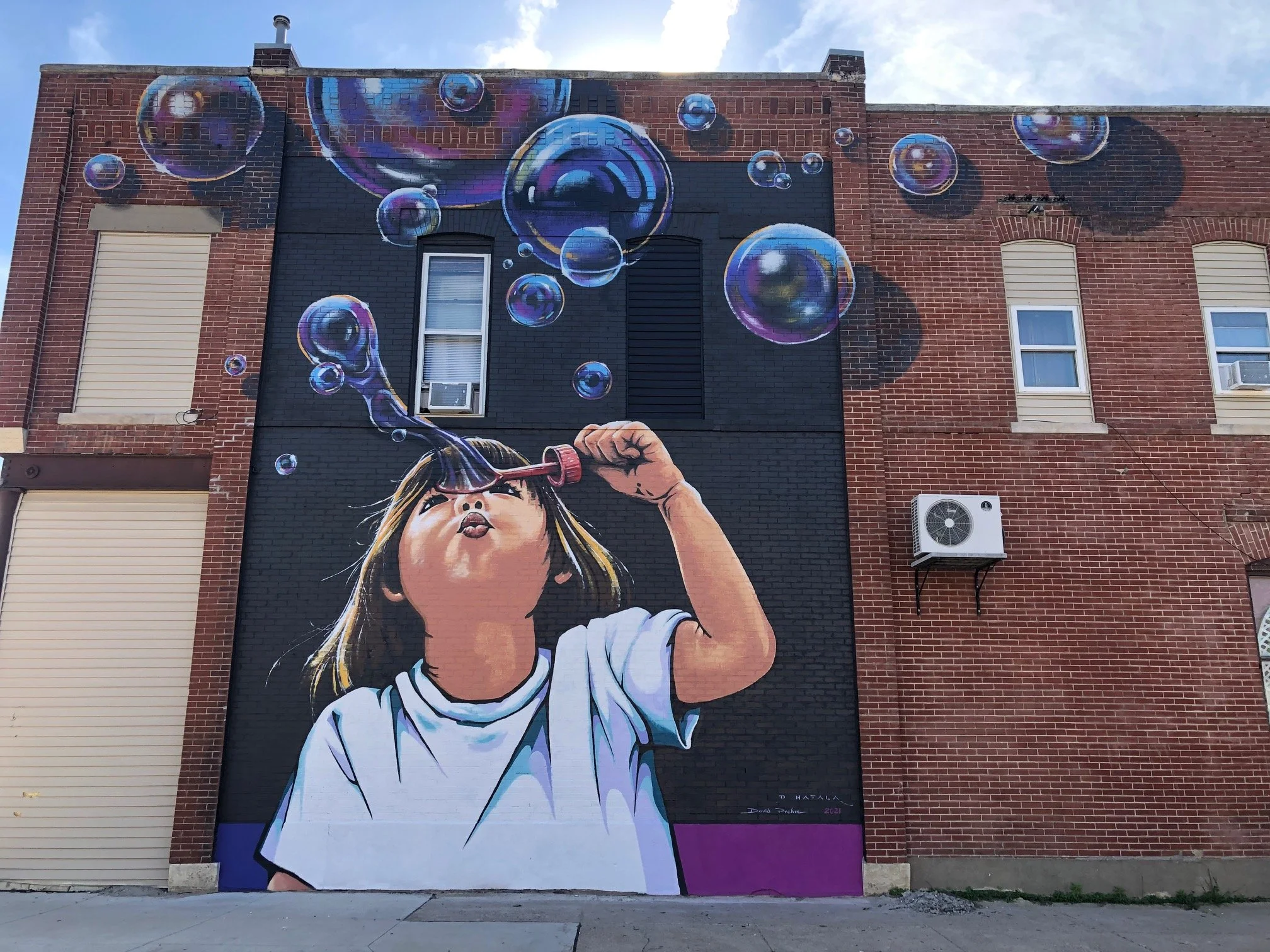 Street art mural of a young girl blowing bubbles into the air on the side of a brick building. The girl is wearing a white t-shirt, and above her, colorful soap bubbles are floating. The background includes red brick and black painted sections on the