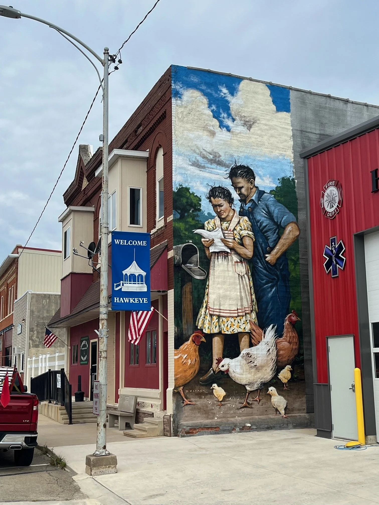 Colorful mural depicting a woman and a man reading a paper, surrounded by chickens, on the side of a building in Hawkeye.