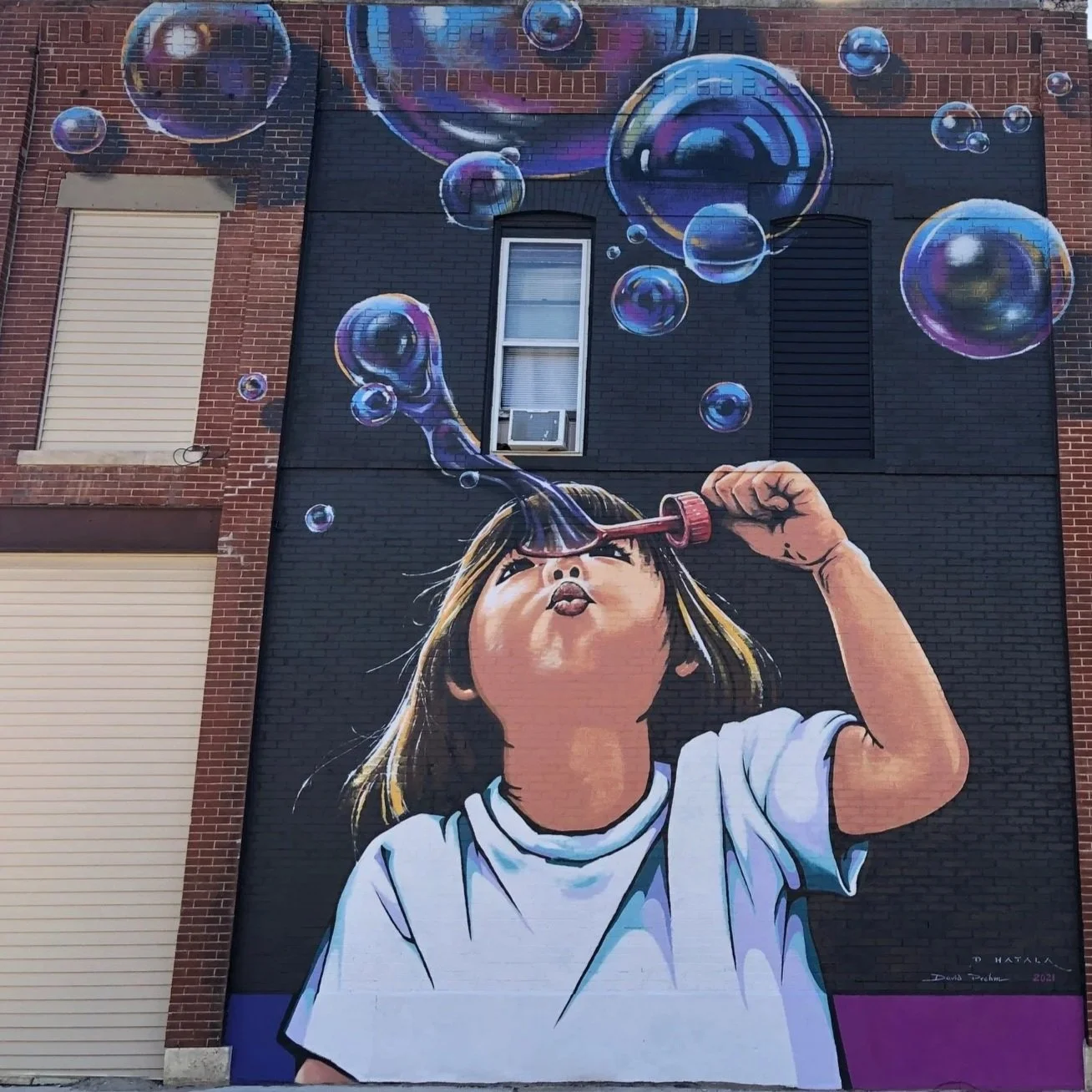 A mural of a young girl blowing bubbles with a bubble wand, with large, colorful bubbles floating around her, painted on the side of a brick building.