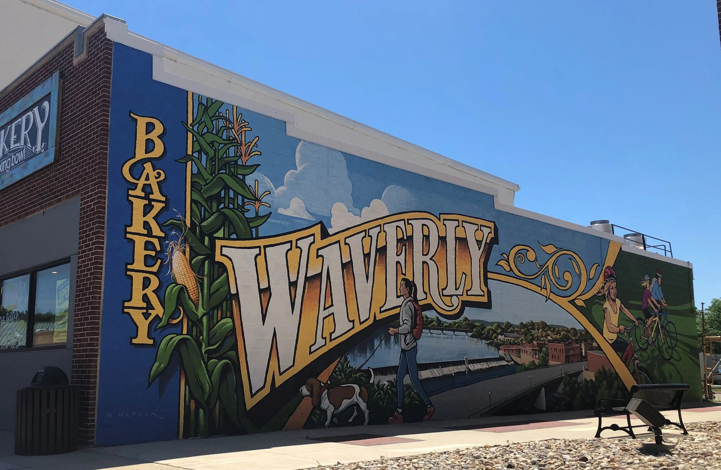 Colorful mural on a building wall with large yellow and white text reading 'Waverly' and 'Bakery'. The mural depicts a woman walking her dog, a corn stalk, and two cyclists riding along a trail, with a river and houses in the background under a blue