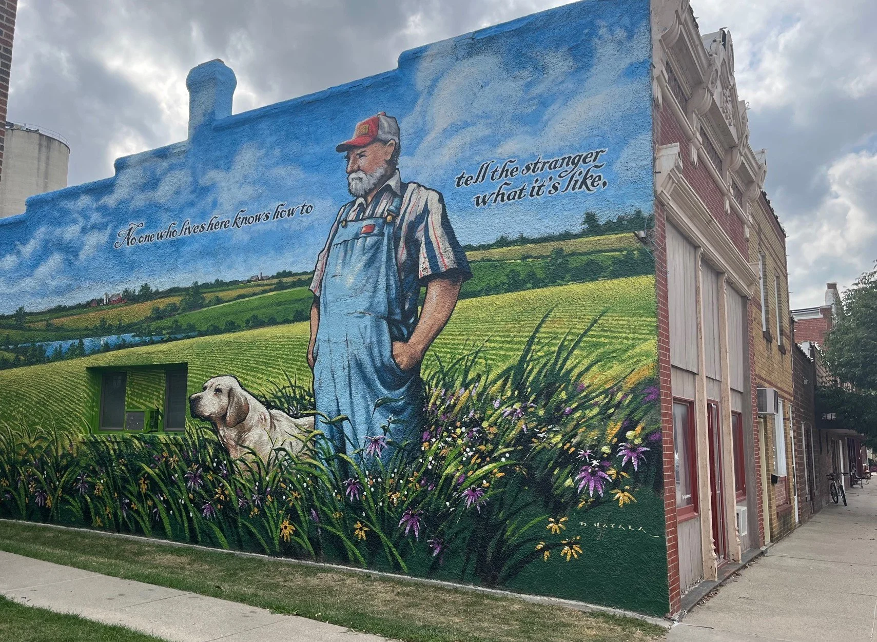 Colorful mural of an elderly farmer with a beard, wearing overalls and a baseball cap, standing in a green field with flowers and a dog beside him. The sky is blue with clouds, and there are inspirational words: "No one who lives here knows how to te