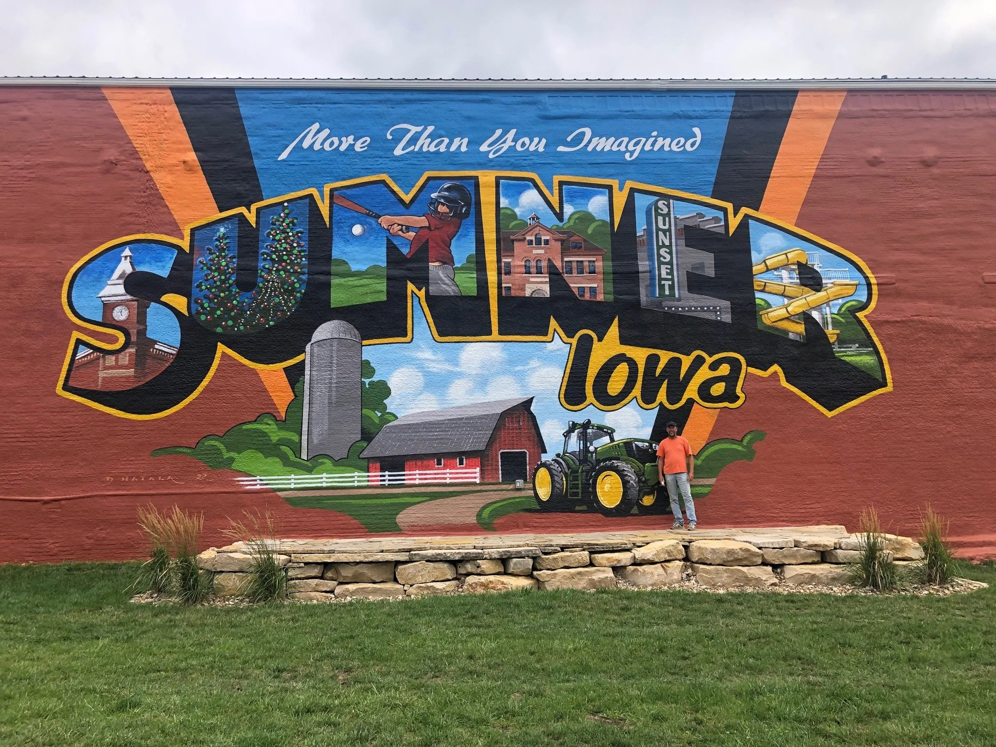 Colorful mural depicting symbols of Iowa, including a clock tower, Christmas trees, a golfer, a historic building, a water slide, a silo, a barn, and a tractor, with the text 'More Than You Imagined' and 'Iowa'.