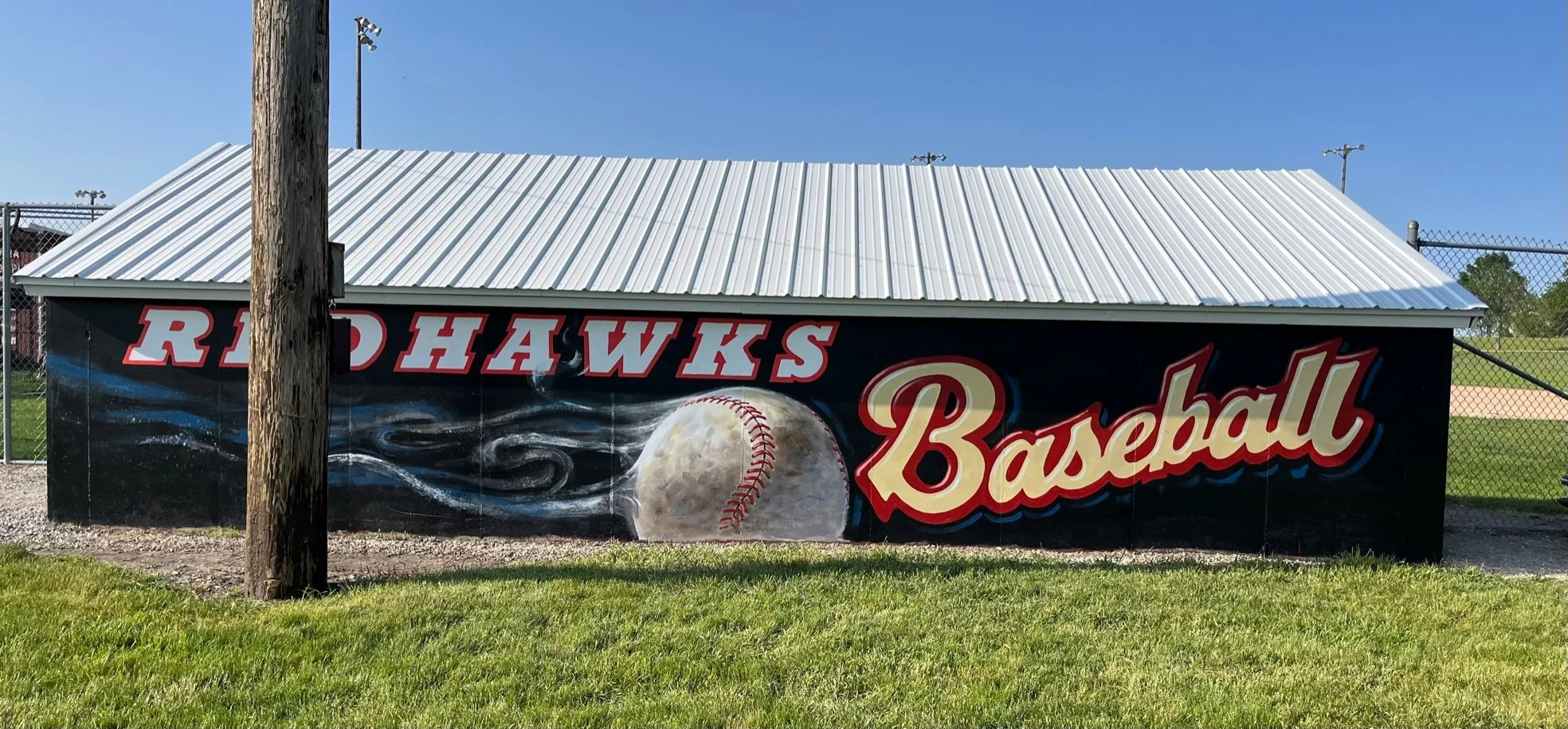 A painted mural on a building featuring the words 'Redhawks Baseball' with a large baseball in the center, spray-painted with a trail effect, against a black background with blue and white accents.