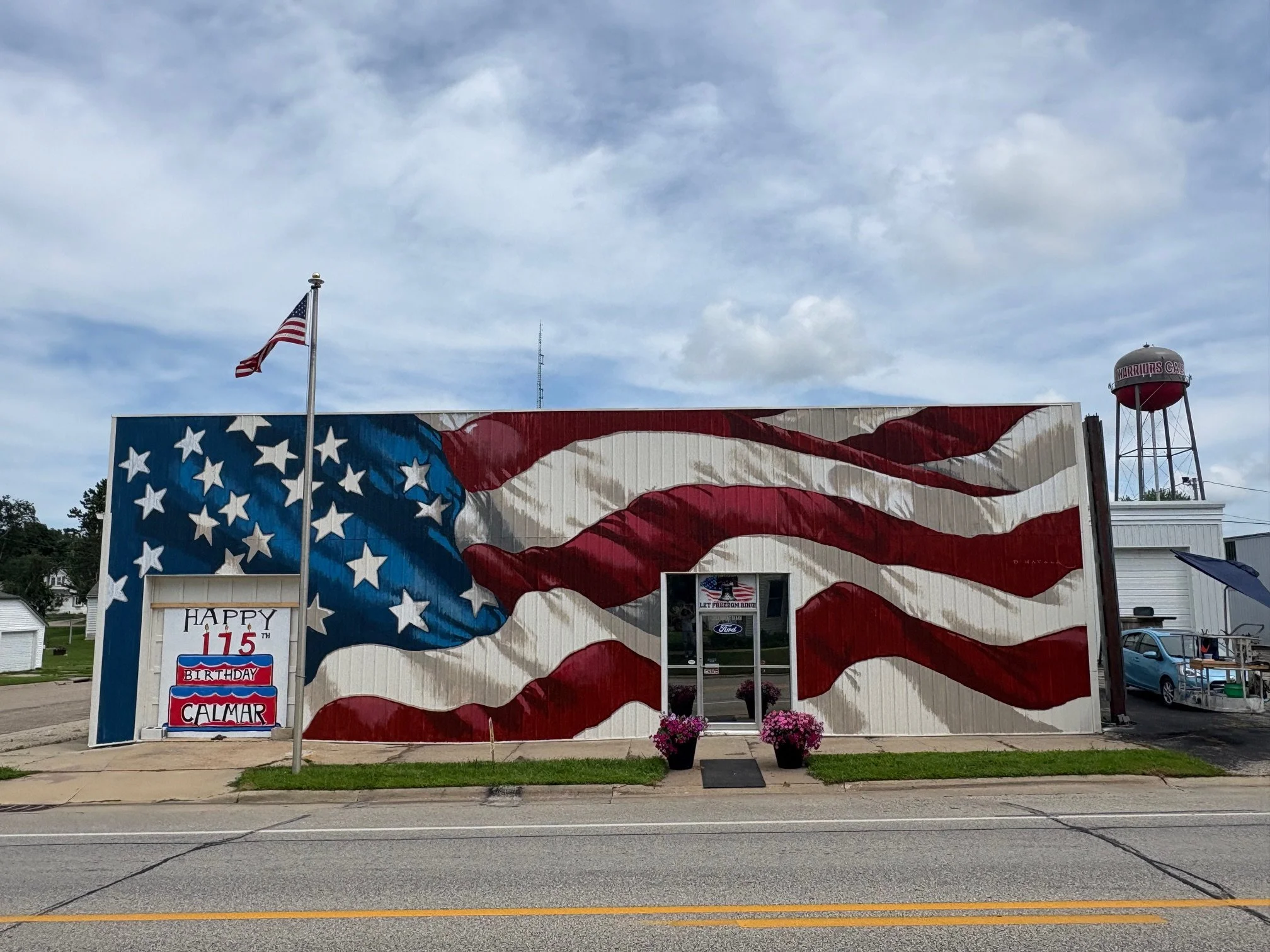 Building painted with an American flag mural, with a flagpole in front, two potted flowers outside the entrance, and a sign celebrating Calmar's 115th birthday.
