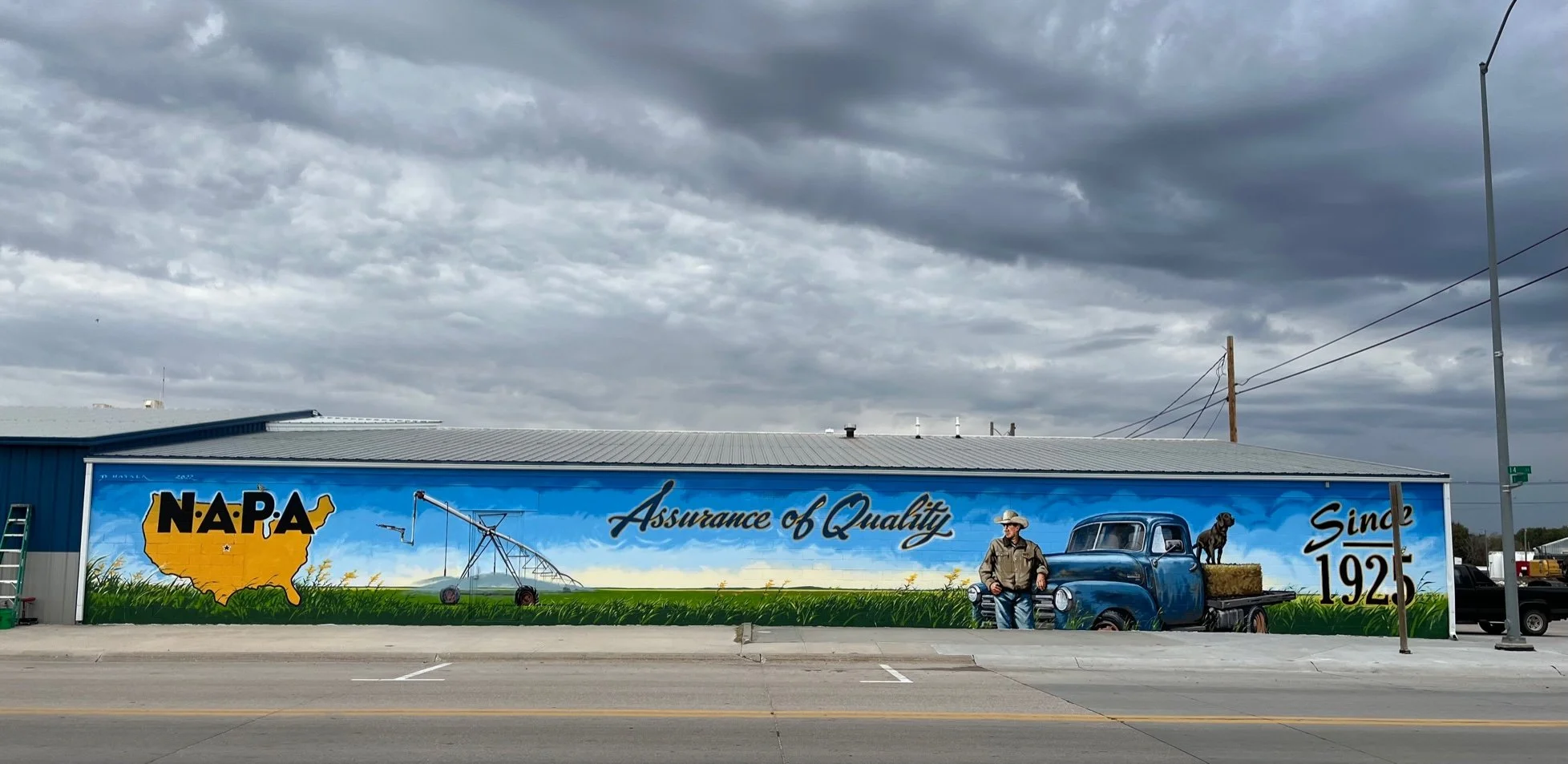 A mural on the side of a building features the NAPA logo with a yellow shape of the United States, a man in cowboy attire, a vintage blue truck with hay on the back, a black dog, and the text 'Assurance of Quality Since 1925' against a grassy landsca