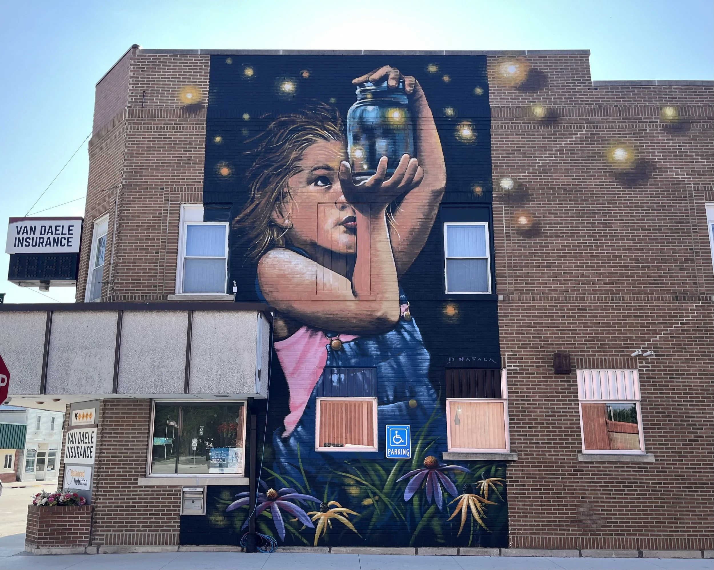 Large mural on brick building depicting a young girl with long hair holding a jar, with lights around her, surrounded by flowers at the base.