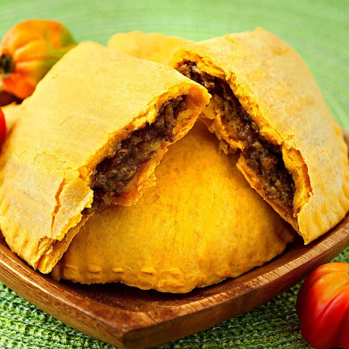 Two sliced, golden-brown empanadas with beef filling in a wooden dish, surrounded by small tomatoes on a green woven placemat.