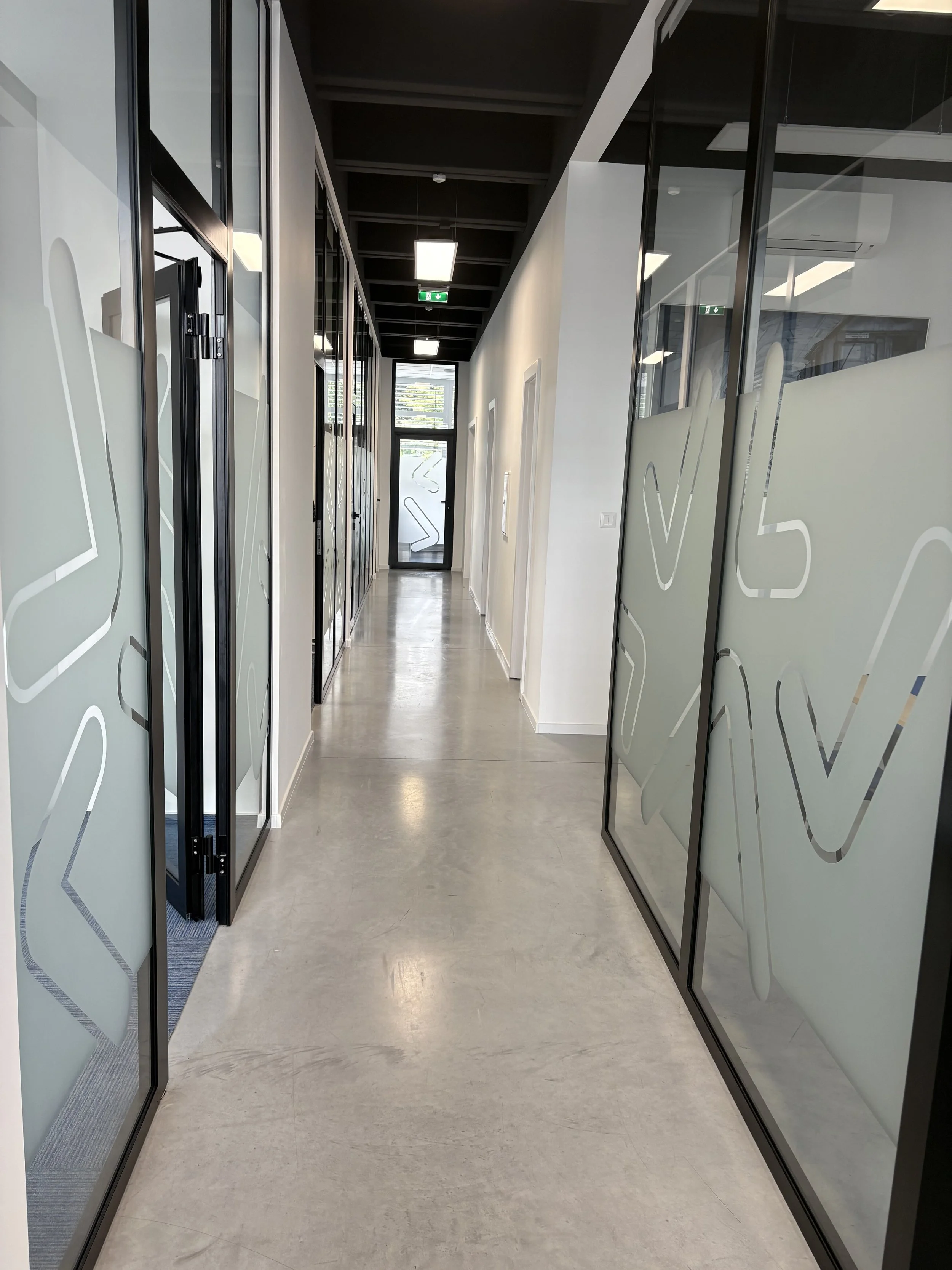 A modern office corridor with glass-walled rooms on both sides, frosted glass designs, white walls, grey flooring, and ceiling lights.