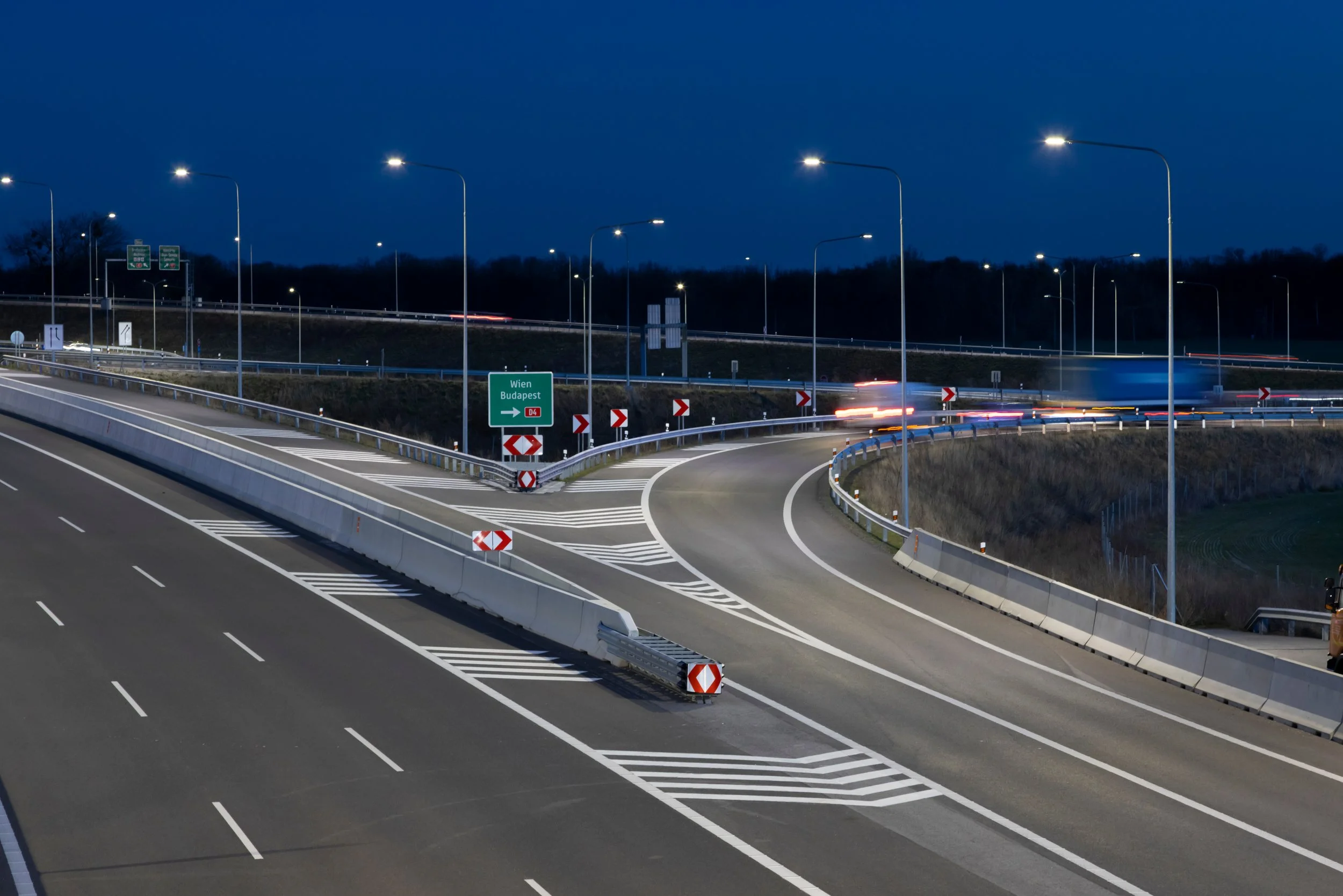 Night view of a multi-lane highway interchange with road signs indicating directions to Wien and Budapest, illuminated streetlights, and blurred vehicle lights.