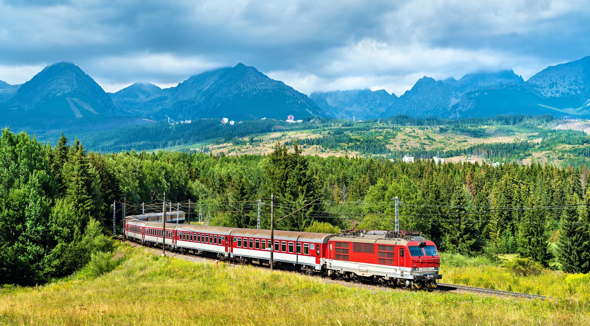A red electric train traveling through a lush green landscape with mountains in the background under cloudy skies.