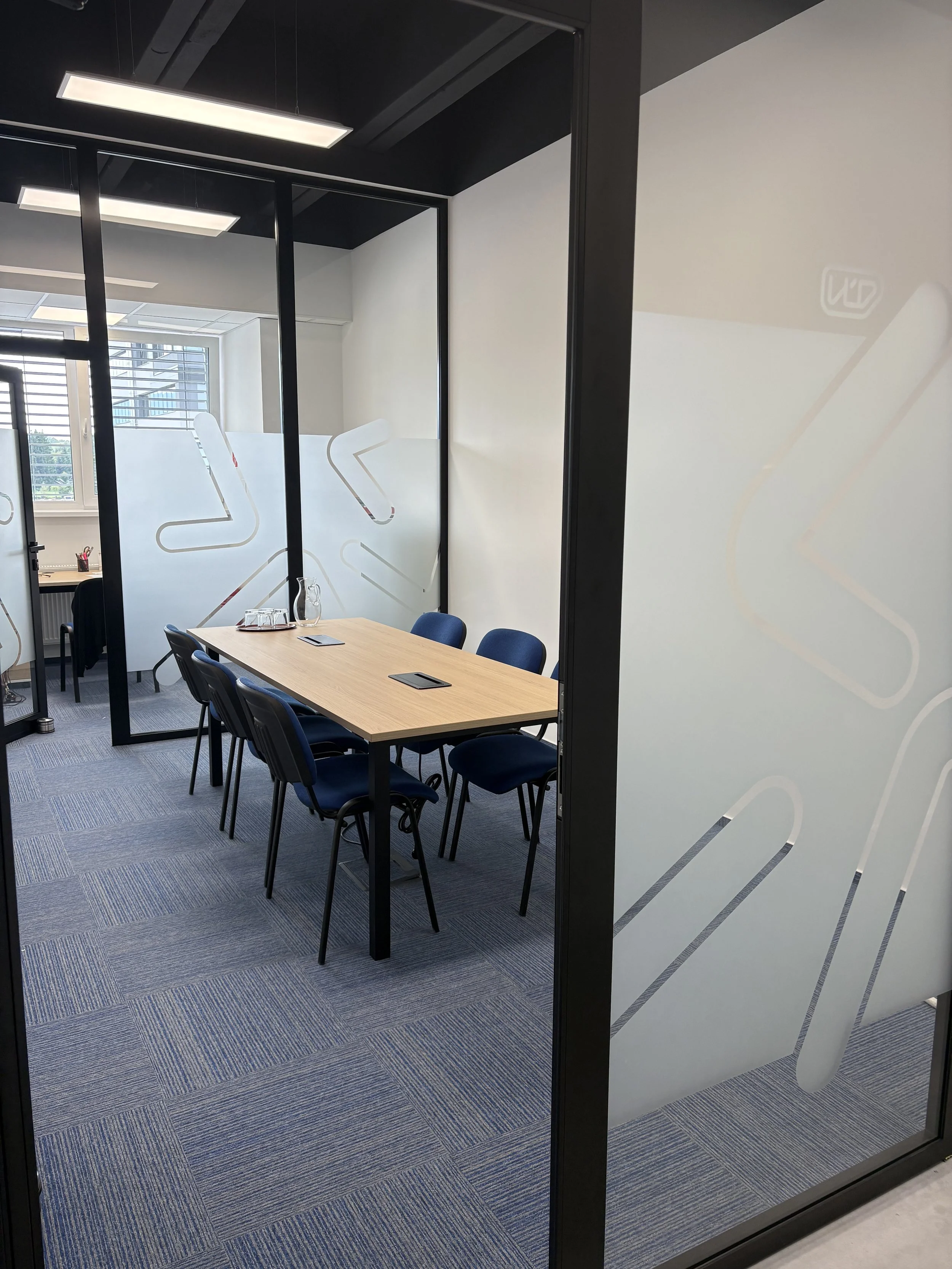 A modern business conference room with a light wood table, six blue chairs, a glass pitcher, glasses, tablet devices, and a window with blinds, enclosed by glass walls with frosted geometric designs.