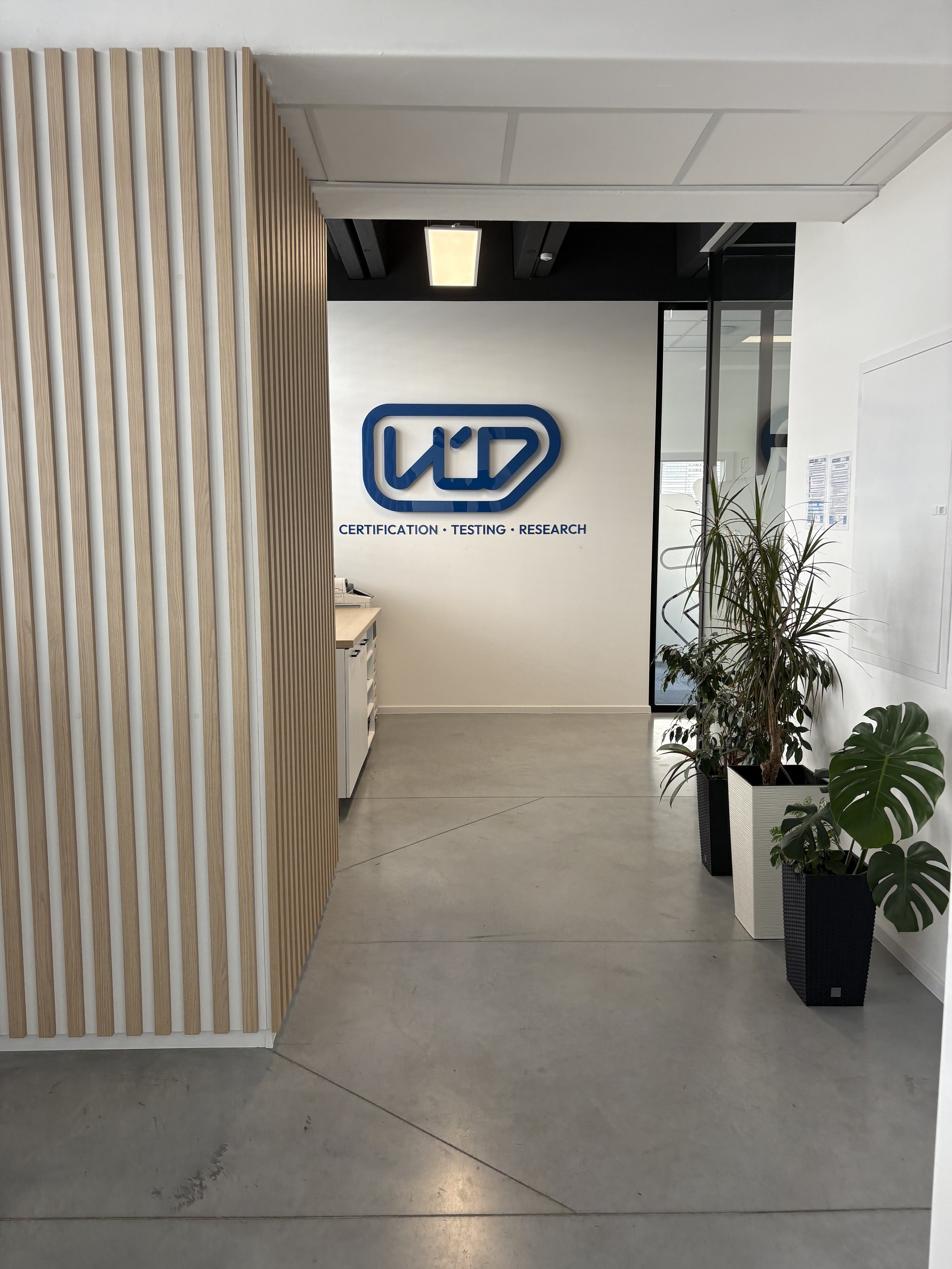 Indoor office space with a wooden slat wall on the left, a white wall with a company logo and text reading 'CERTIFICATION · TESTING · RESEARCH' in blue, potted plants on the right, and a gray concrete floor.