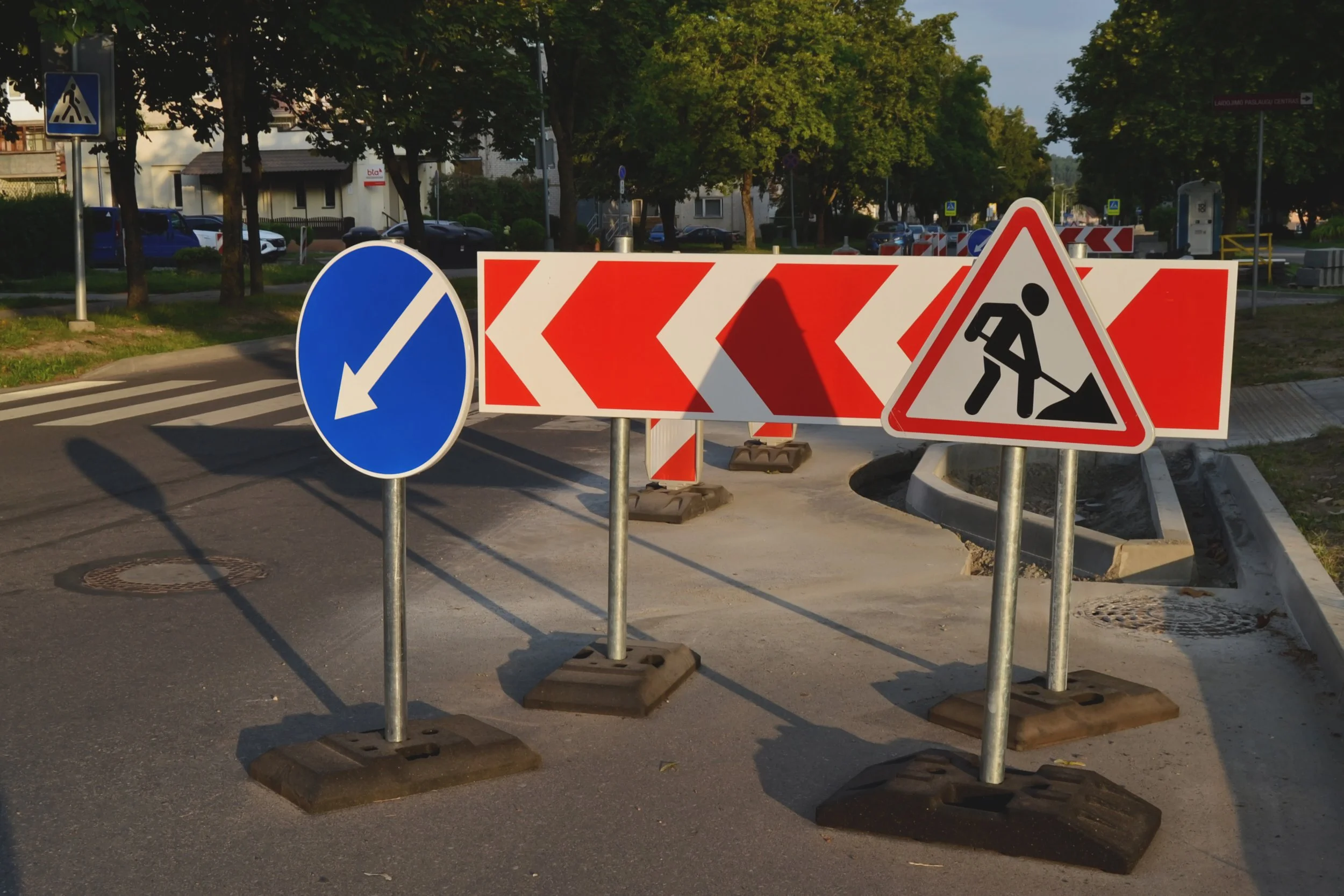 Road construction zone with warning signs including a blue circle with a white arrow pointing left, red and white striped barriers, and a triangular sign indicating road work.