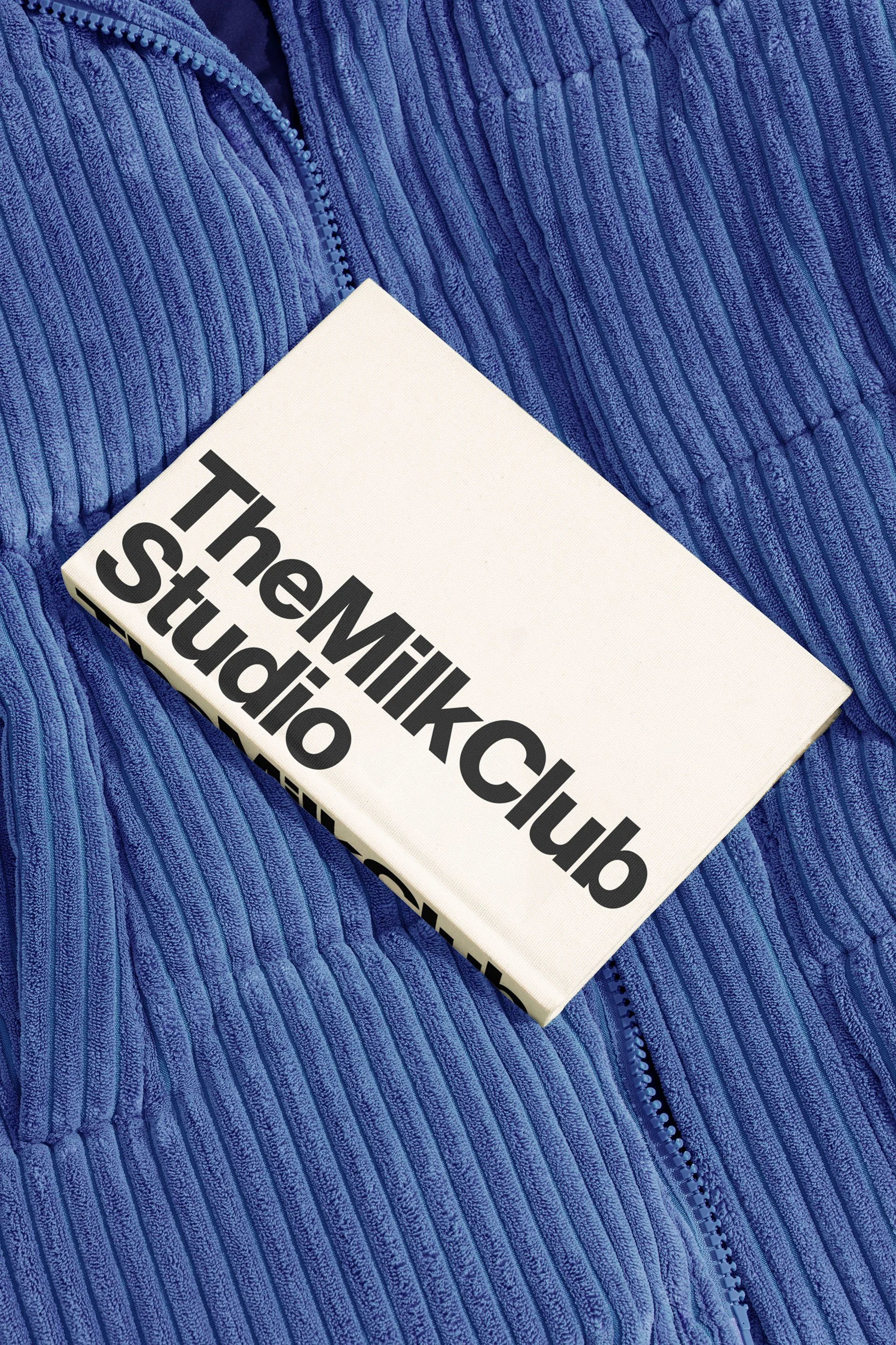 A white rectangular object with the text 'The Milo Club Studio' in bold black letters, placed on a blue corduroy fabric surface.