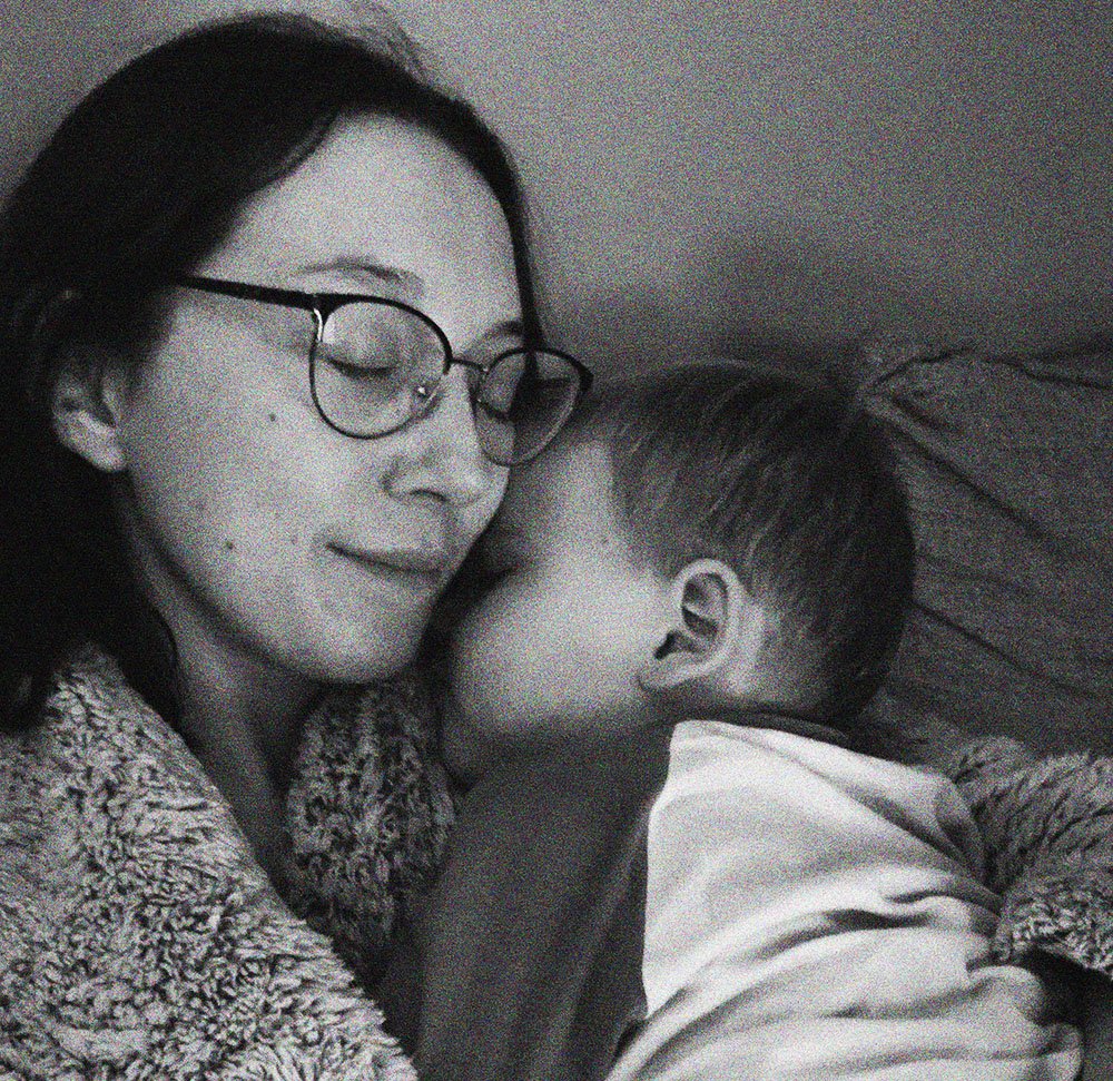 A woman with glasses and dark hair with closed eyes, smiling, cuddling a young boy with short hair, lying on a pillow.