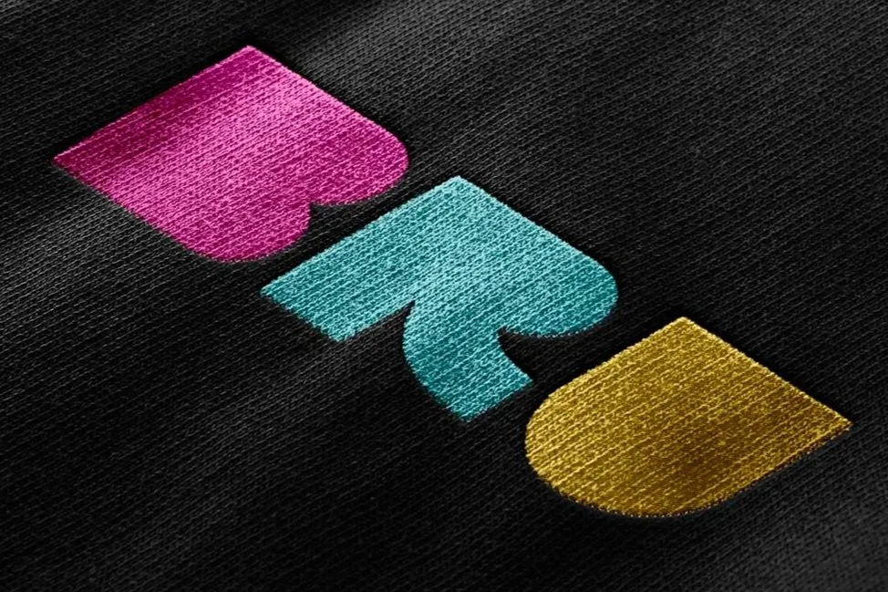 Close-up of embroidered geometric shapes in pink, teal, and gold on black fabric.