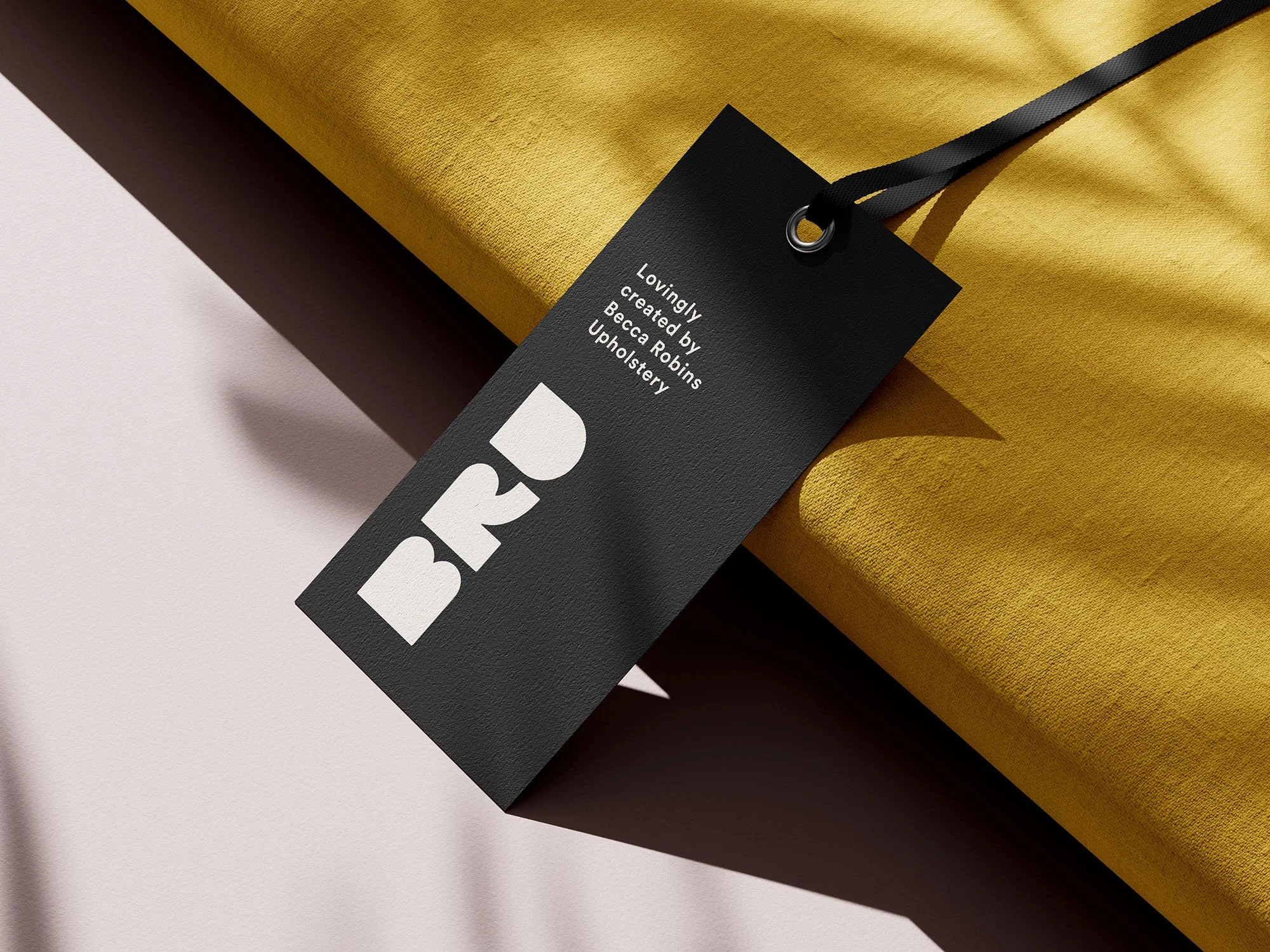 Close-up of a black tag attached to a yellow package. The tag reads 'BR' and states it was created by Becca Robins Upholstery.