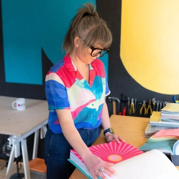 Woman with glasses and a colorful shirt looking at a colorful notebook on a wooden table in a creative workspace.