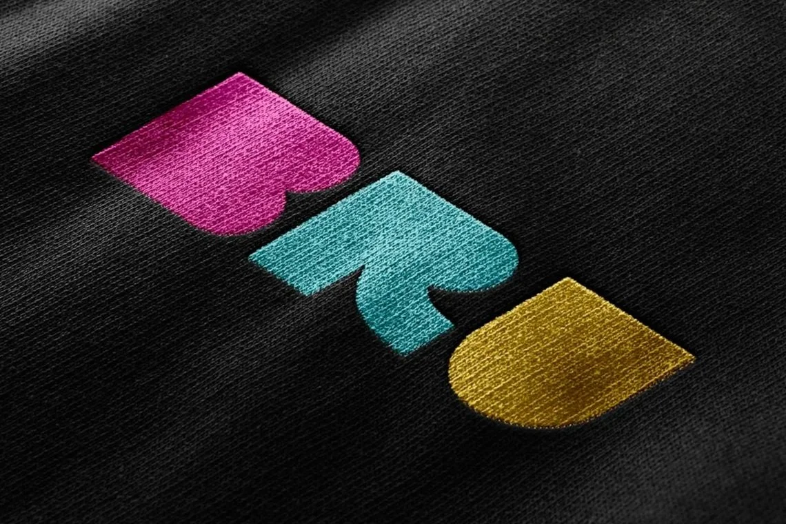 Colorful logo with stylized shapes in pink, blue, and gold on black fabric background.