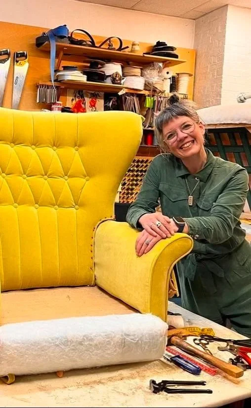 A person smiling and leaning on a yellow upholstered armchair in a workshop or furniture repair space, with tools and supplies on a table and shelves in the background.