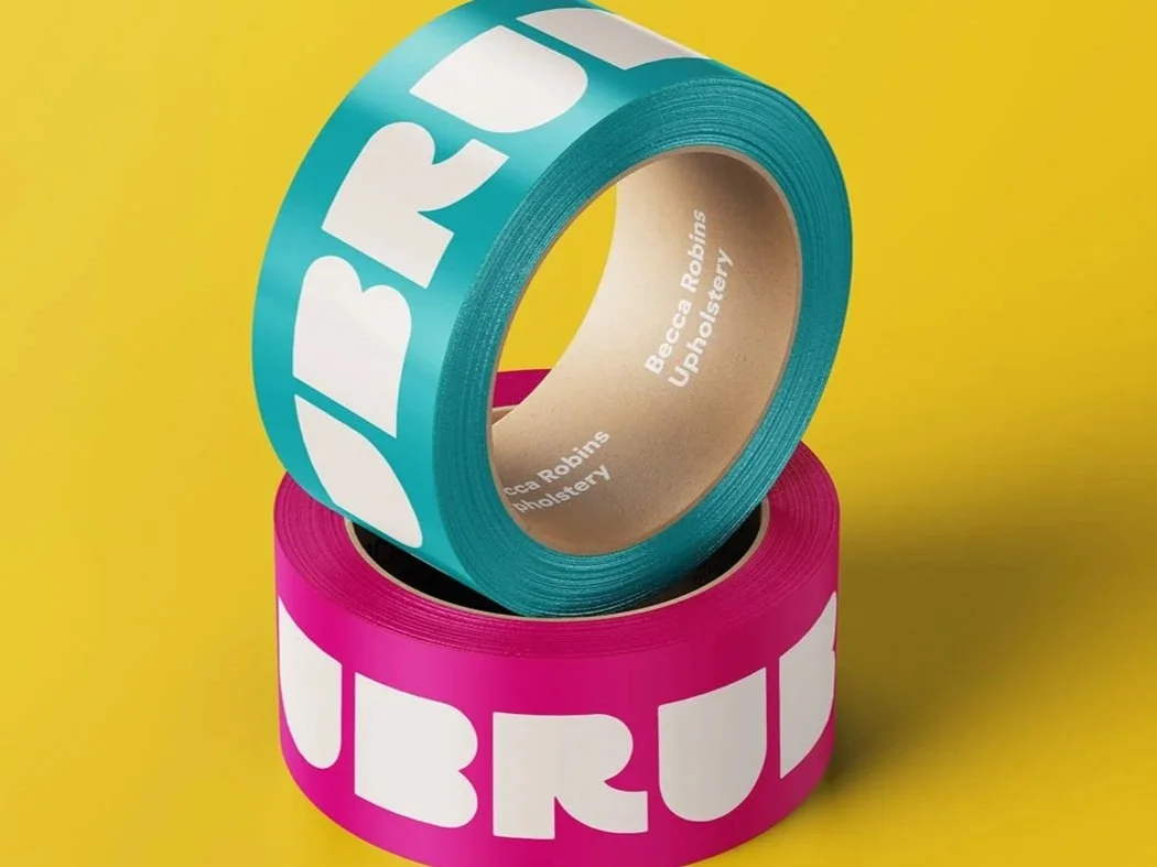 Two rolls of tape, one turquoise with large white letters spelling 'UBRU,' and one pink with large white letters, placed on a yellow background.