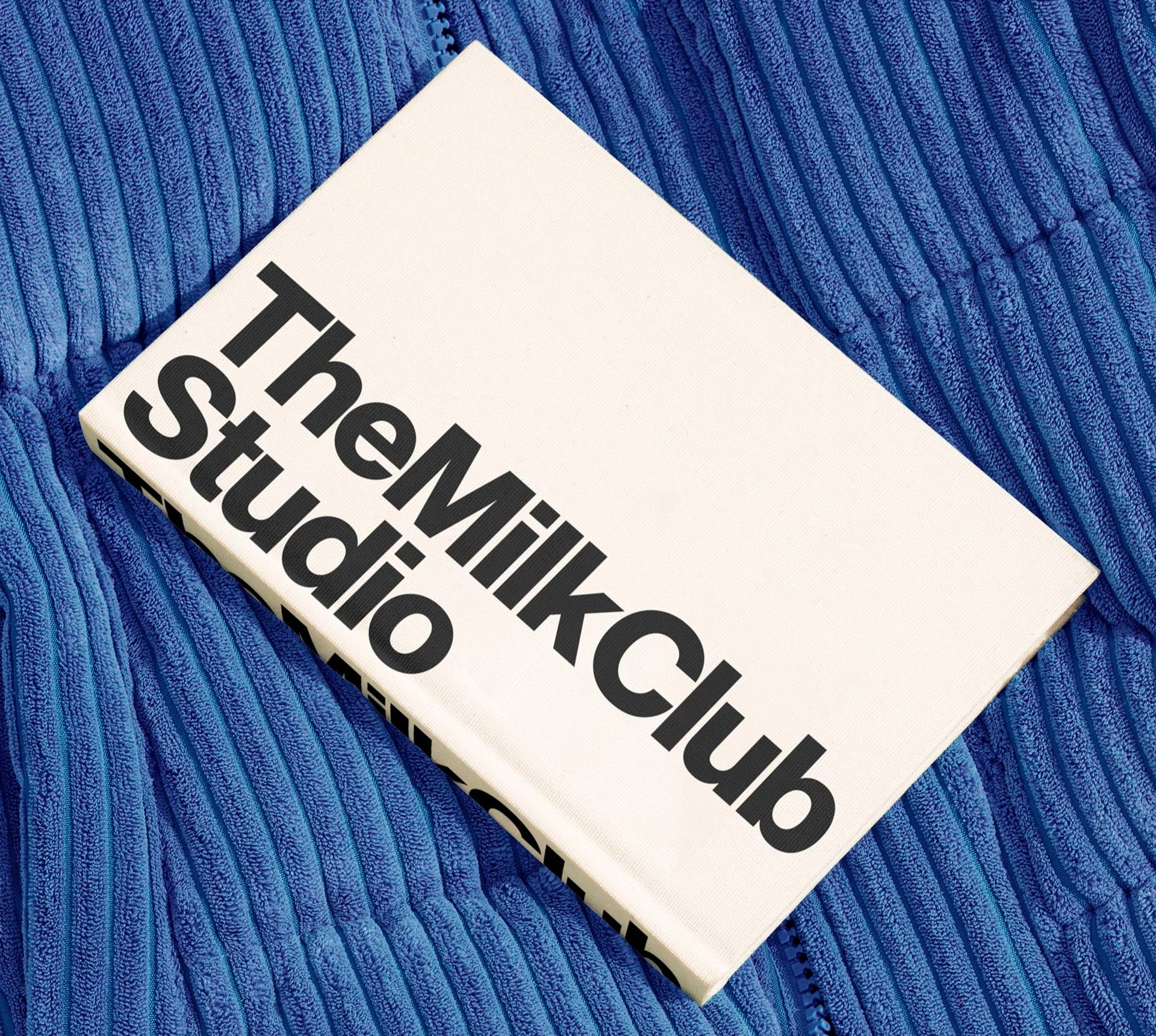 Book titled 'The Milk Club Studio' resting on a textured blue surface.